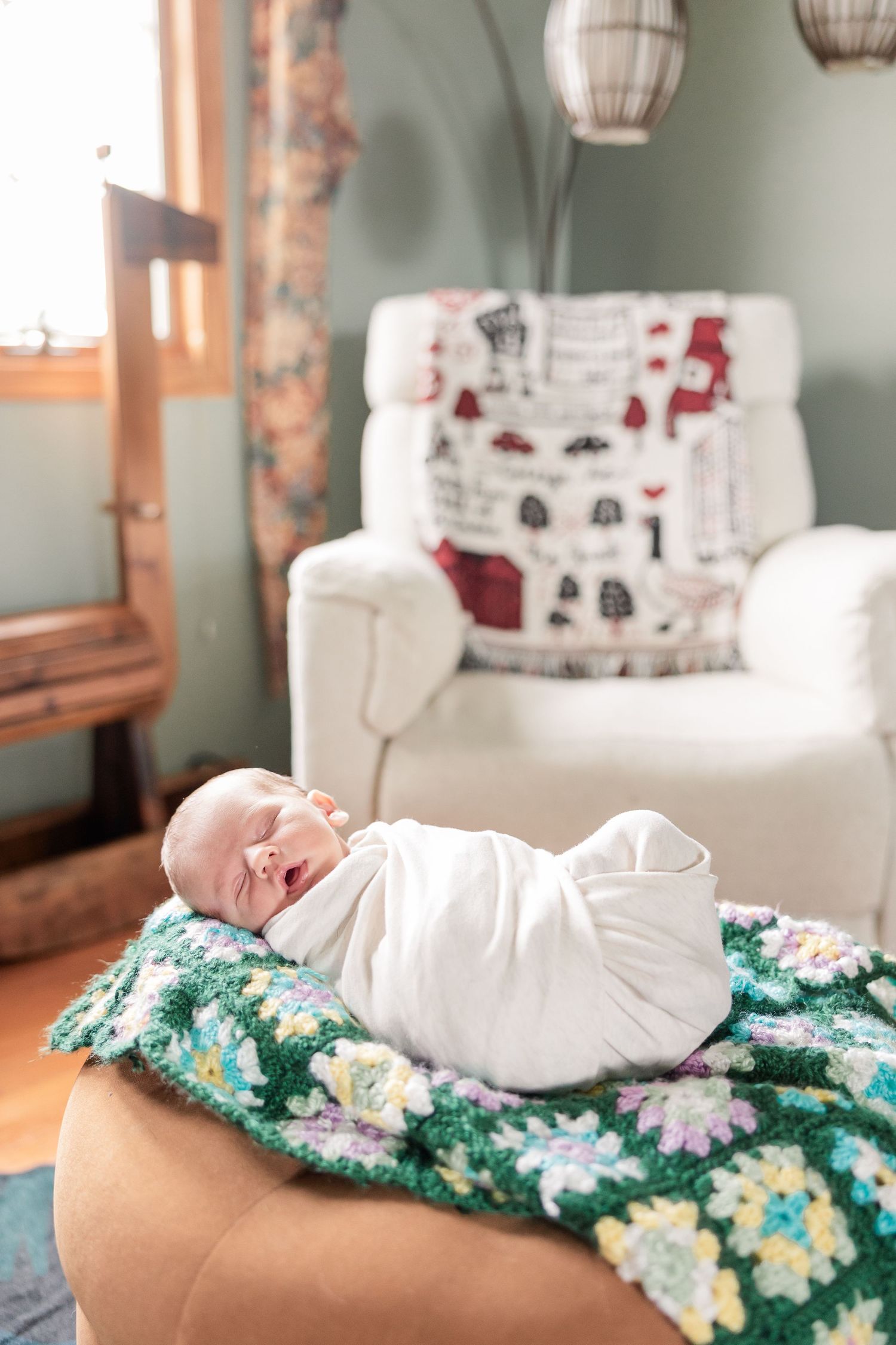 Swaddled newborn baby rests on colorful crocheted blanket on ottoman in cozy nursery room.