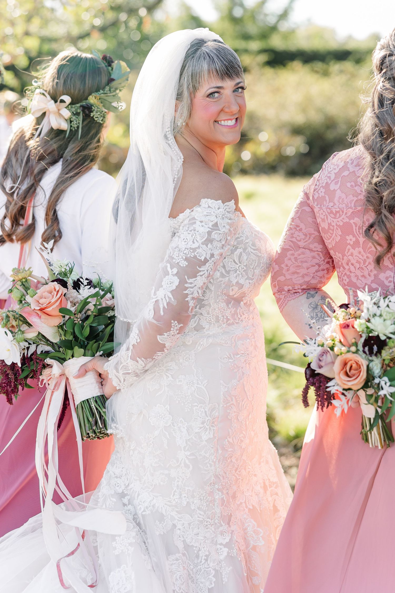 Smiling bride in a lace off-the-shoulder wedding gown and veil poses outdoors with bridesmaids in pink dresses holding floral bouquets.