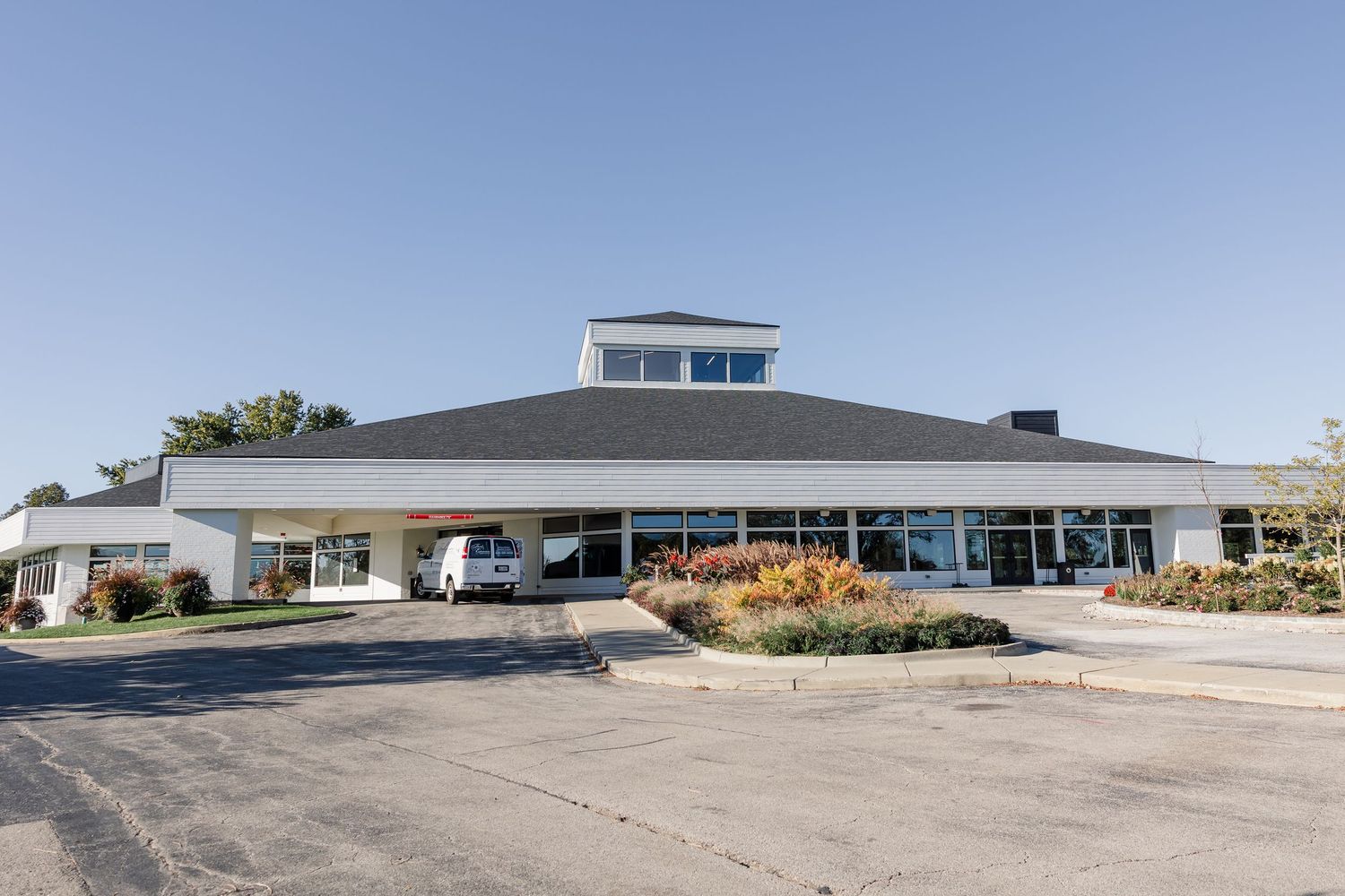 Single-story commercial building with a dark pitched roof, large glass windows, and a parking lot under a clear blue sky.