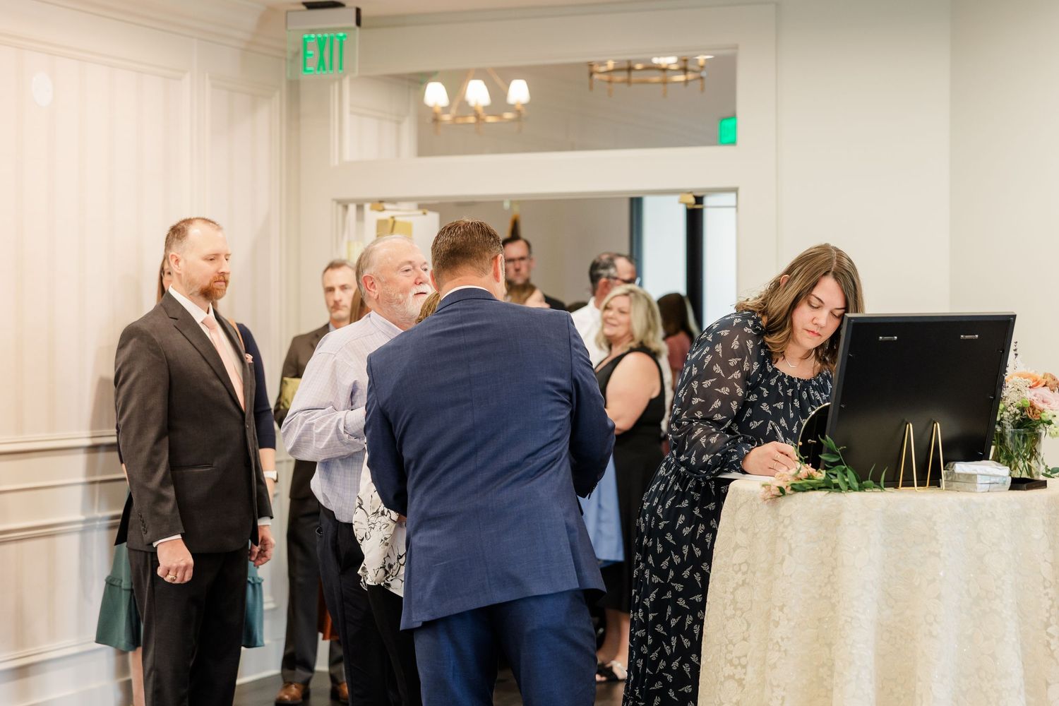Guests in formal attire mingle at an indoor event while a woman in a floral dress writes at a check-in table with a laptop and flowers.
