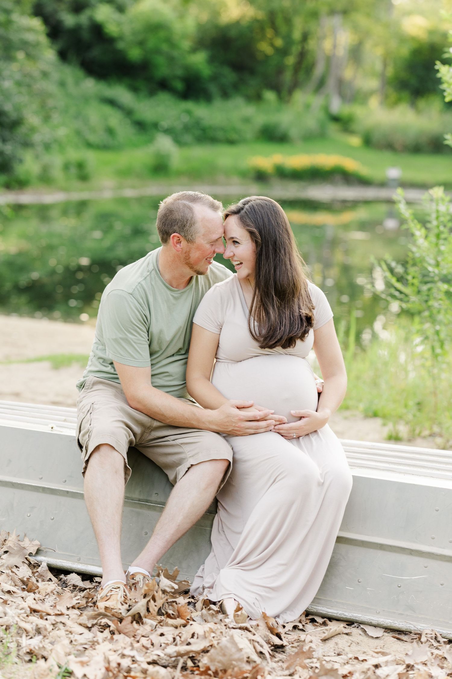 An expectant mother in a flowing white dress poses serenely beside a boat in a tranquil lakeside setting amid summer greenery.