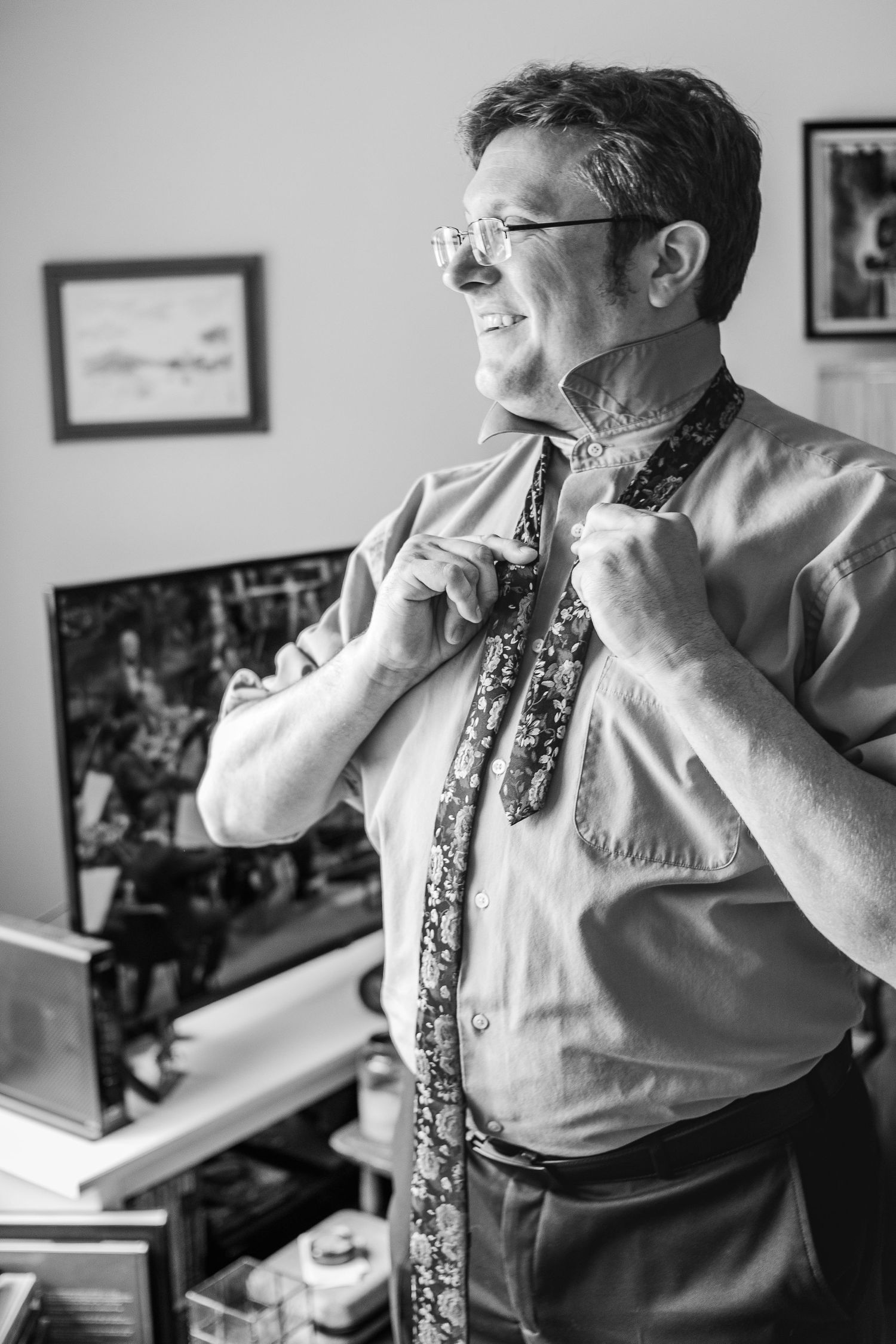 Black and white photo of someone adjusting their tie while wearing glasses and a dress shirt.