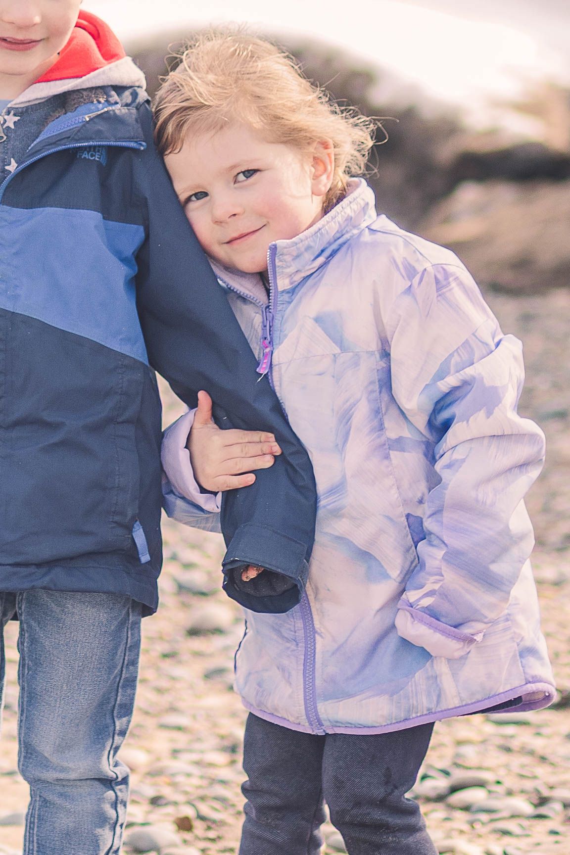 Child in lavender winter jacket smiles while standing on rocky beach.