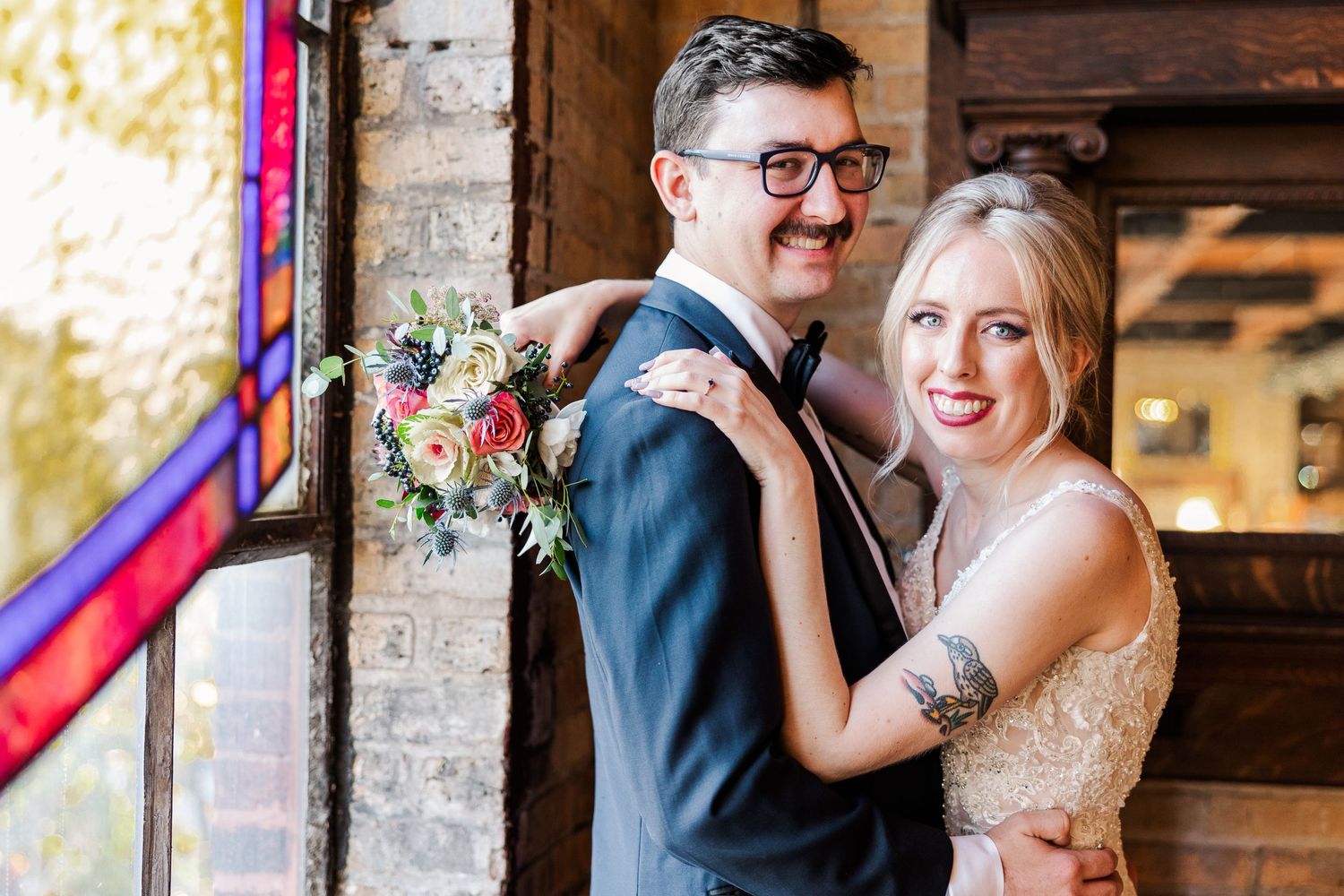 A couple shares a tender moment near a stained glass window in an industrial wedding venue.