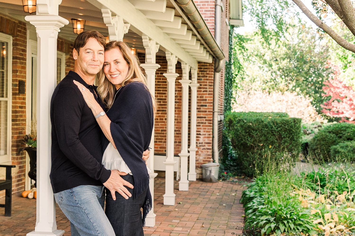 Romantic couple pose beneath white columns on brick patio surrounded by gardens.