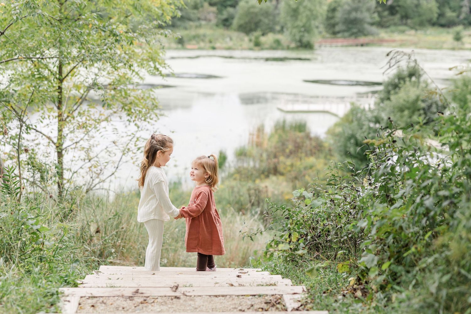 Two young girls stand on wooden steps near a serene lake surrounded by lush greenery in a natural outdoor setting.