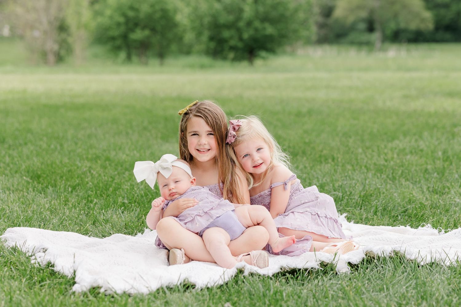 Three young sisters in matching lavender outfits sit together smiling on a white blanket in a lush green park.