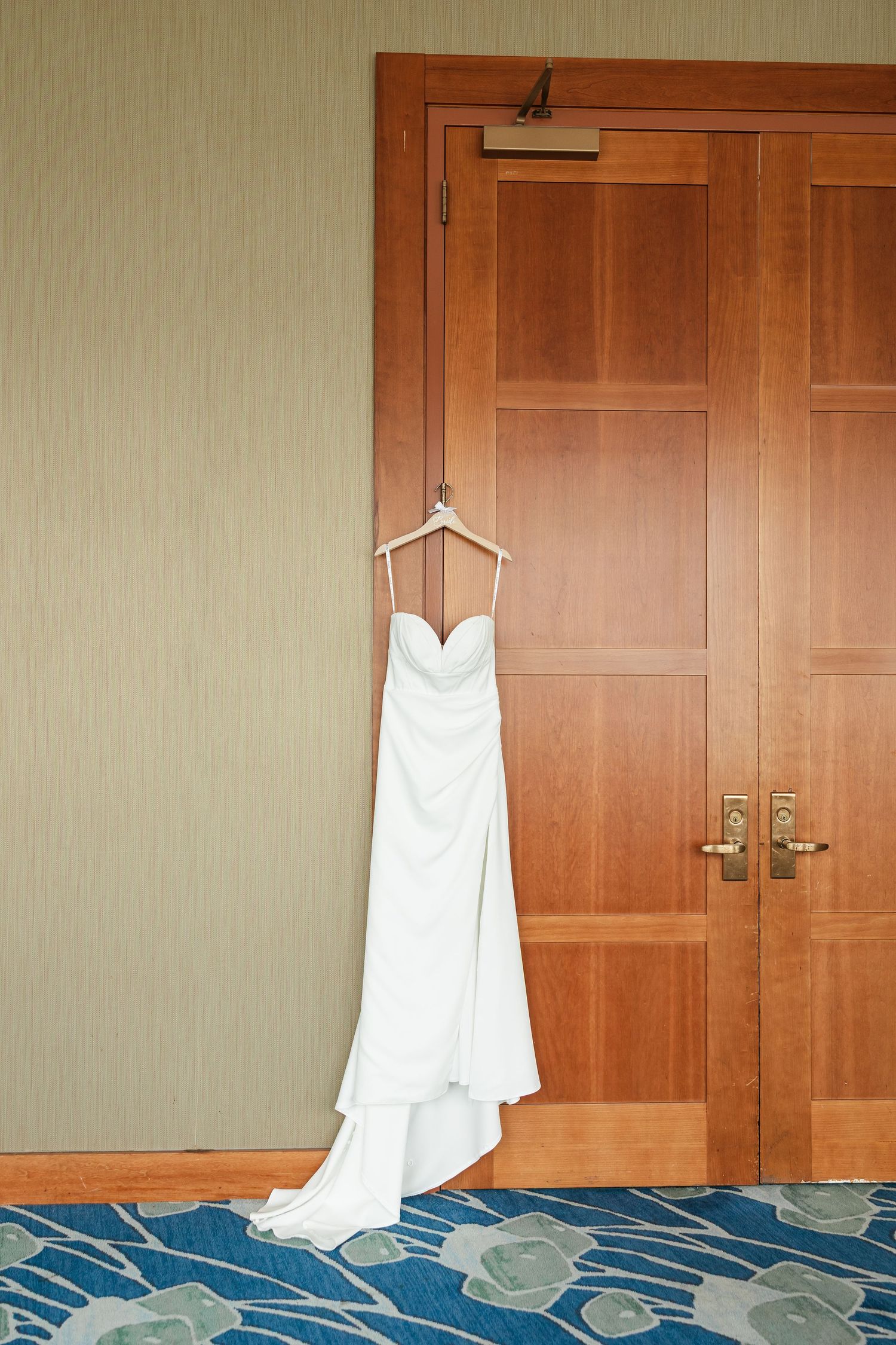 White strapless wedding dress hanging on wooden door in hotel room with blue floral carpet.