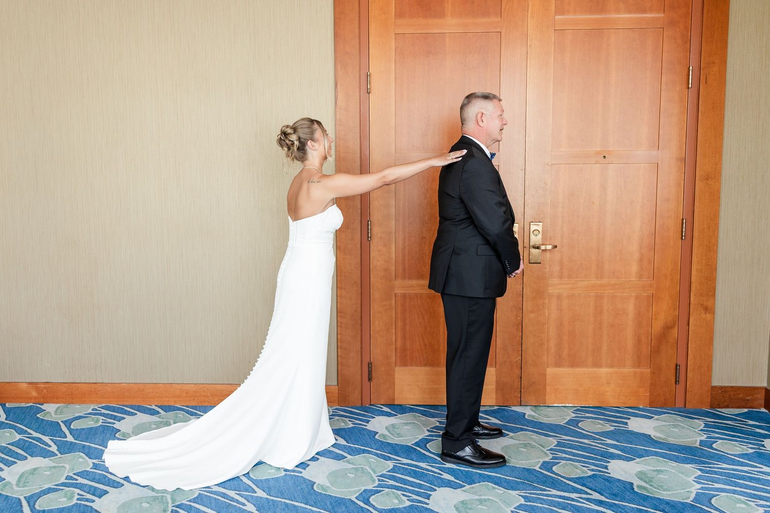 Bride in white gown tapping groom on shoulder for first look moment in hotel hallway.