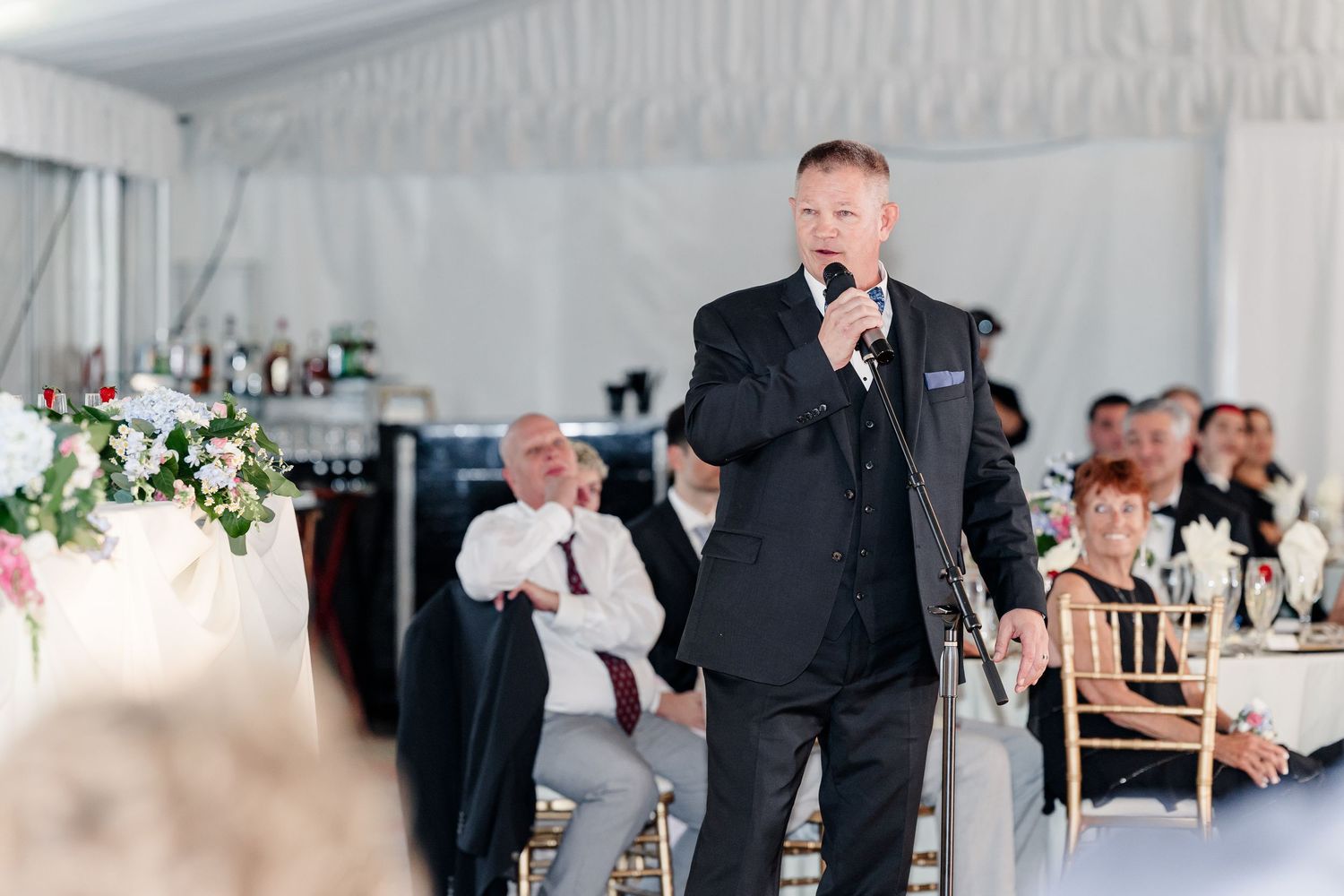 Man in black suit speaking into microphone during wedding reception speech.