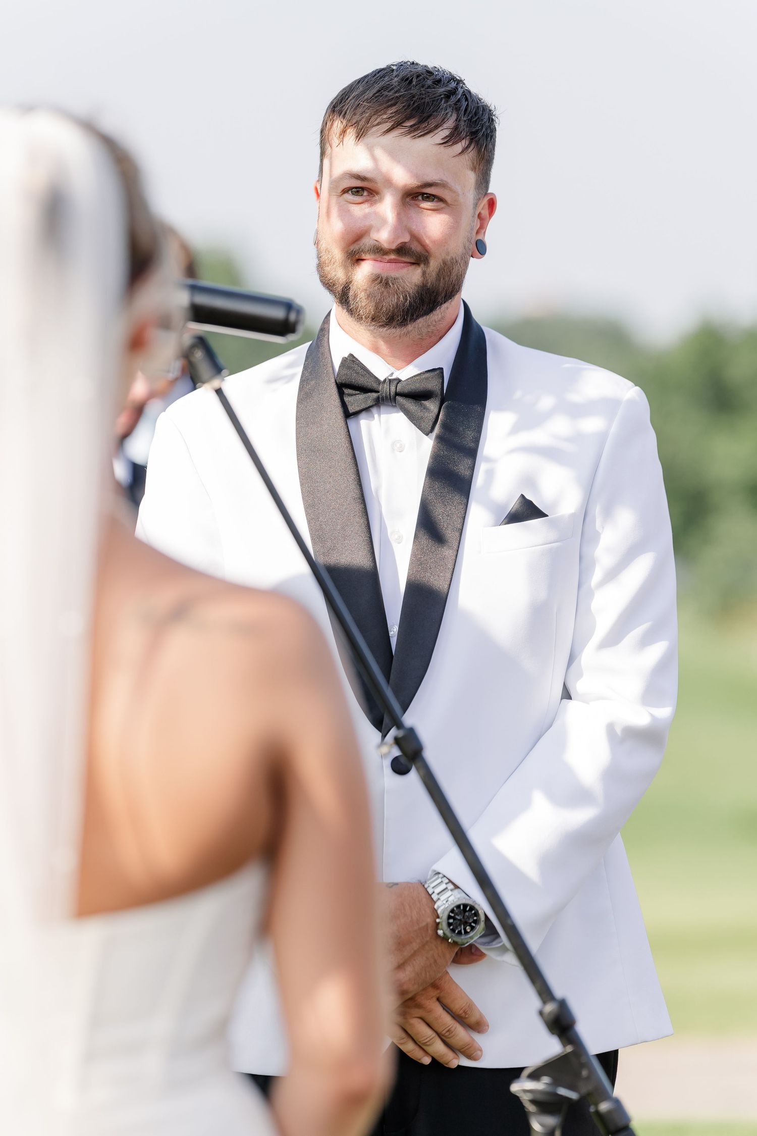 Groom in white tuxedo smiling at bride during outdoor wedding ceremony.