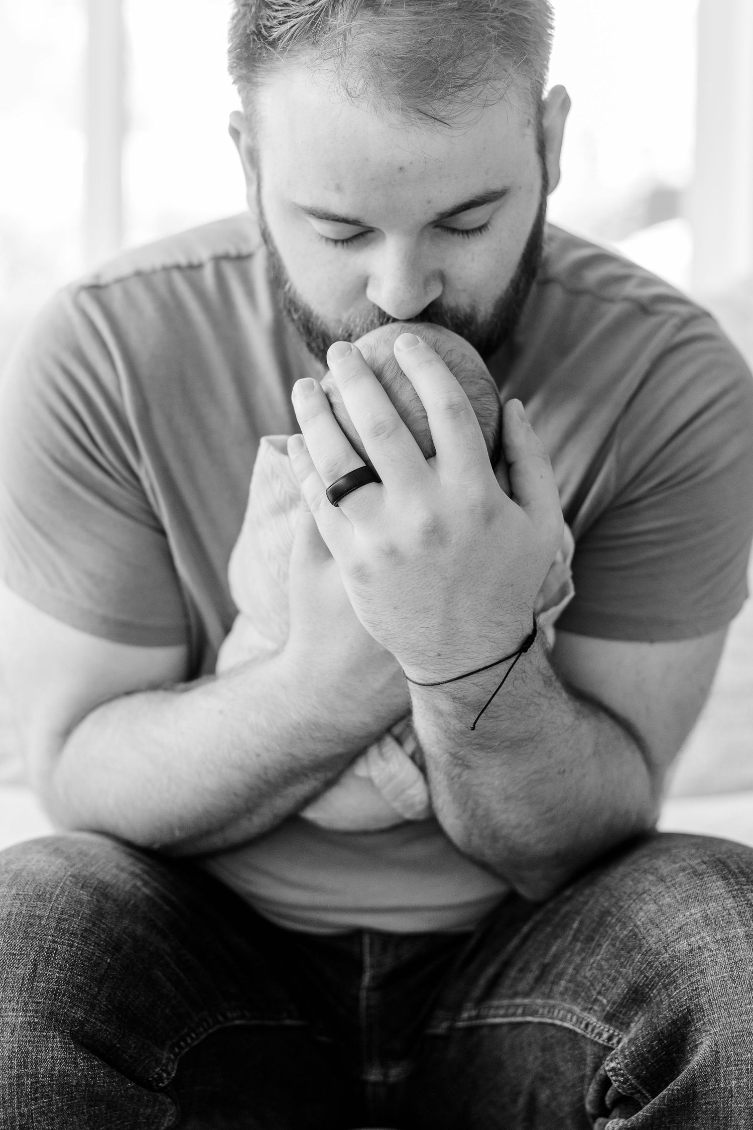 Bearded man in gray t-shirt kissing a newborn baby while seated, black and white portrait photo.