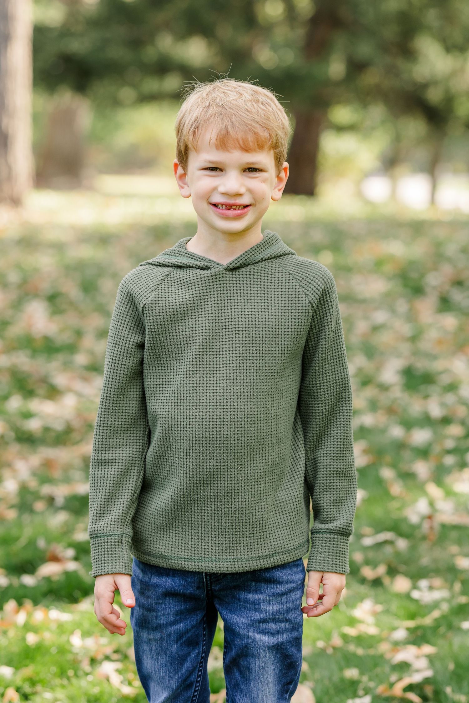 Smiling redhead boy in green waffle hoodie and jeans posing outdoors in a leaf-covered park.