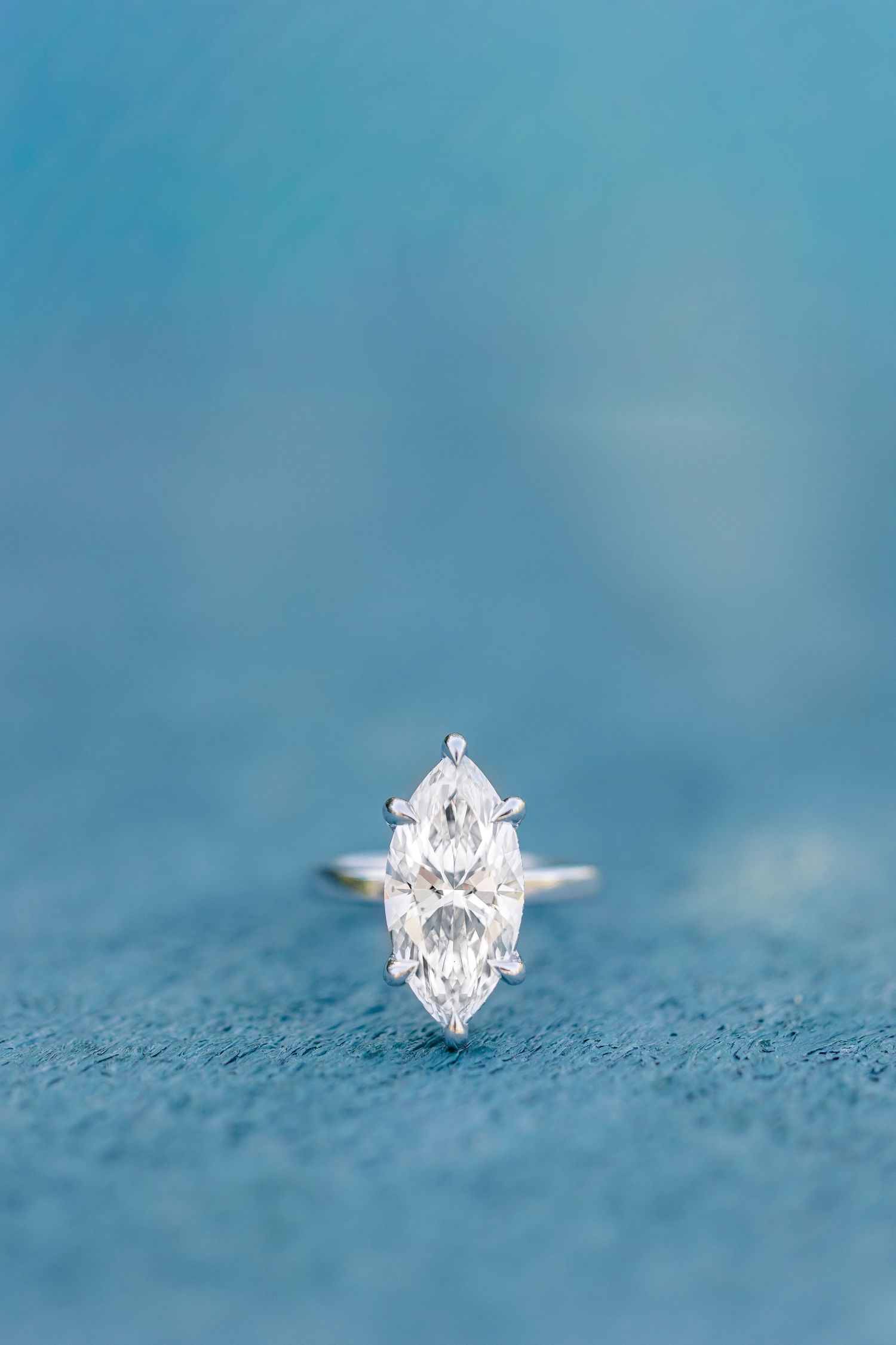 Elegant marquise cut solitaire diamond engagement ring displayed on blue textured surface with silver band.