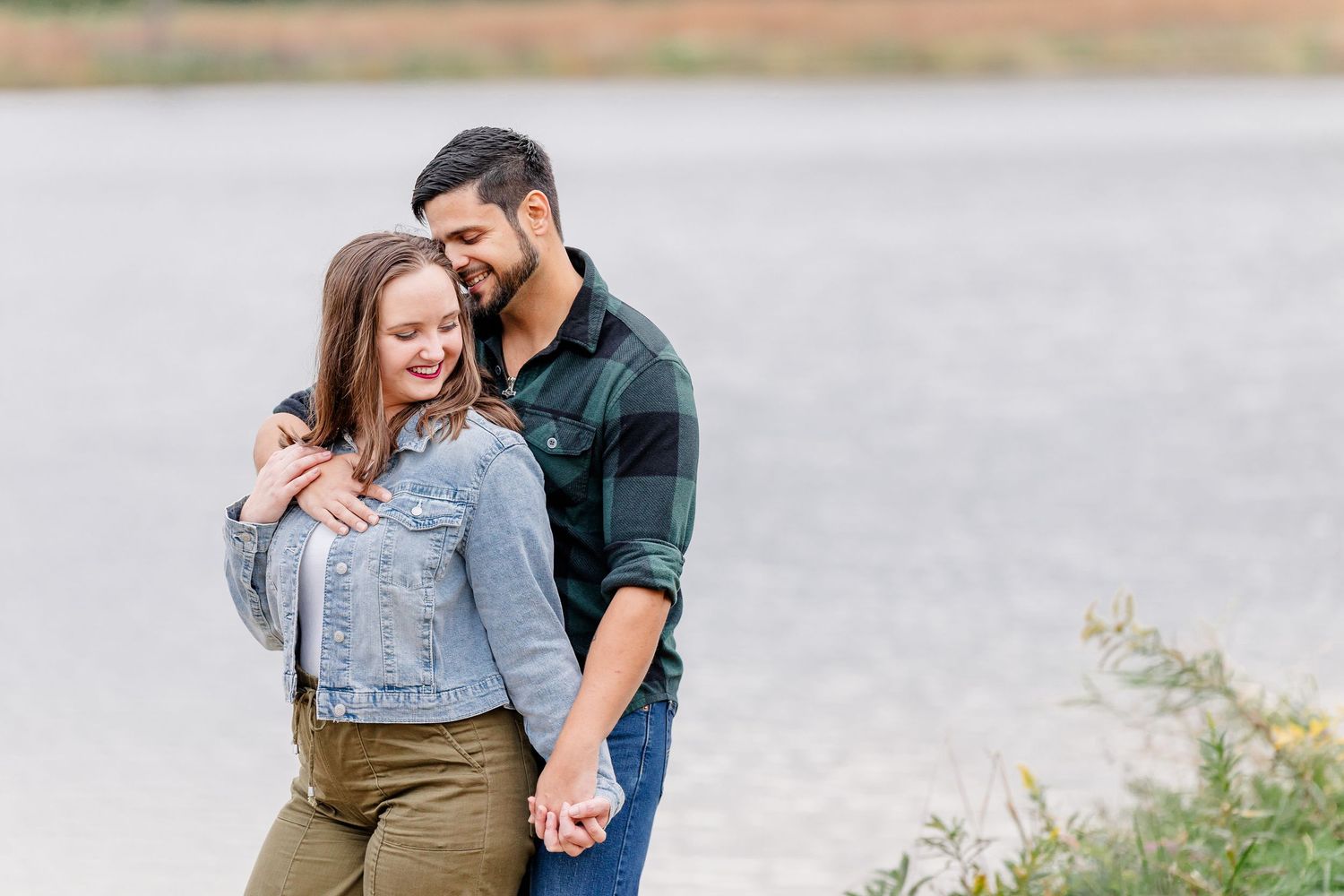 Couple in denim jacket and green plaid shirt embracing and holding hands by a lake during an engagement photo session.
