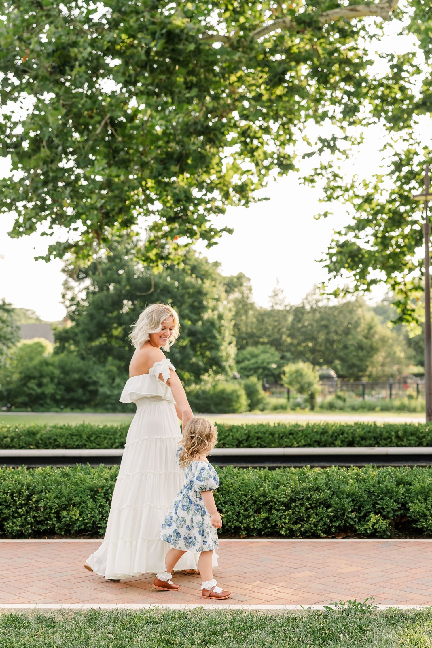 A mother in a white dress dances with her young daughter in a floral outfit on a brick path surrounded by lush greenery.
