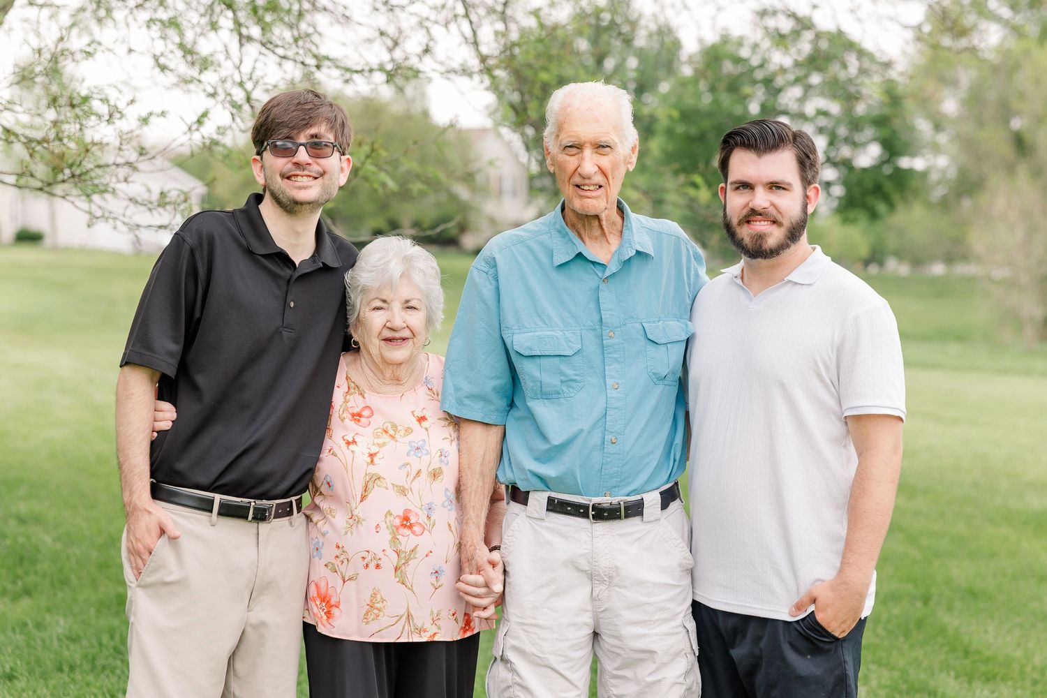 Four family members, two elderly and two young adults, smiling outdoors on green grass with trees in background.