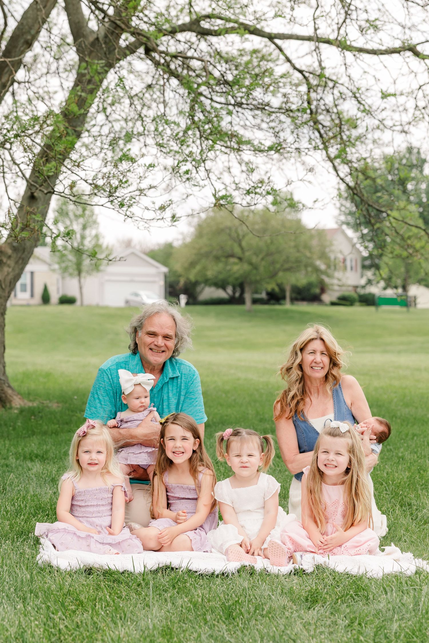 Grandparents with six young granddaughters sitting on blanket under spring trees during family portrait session.