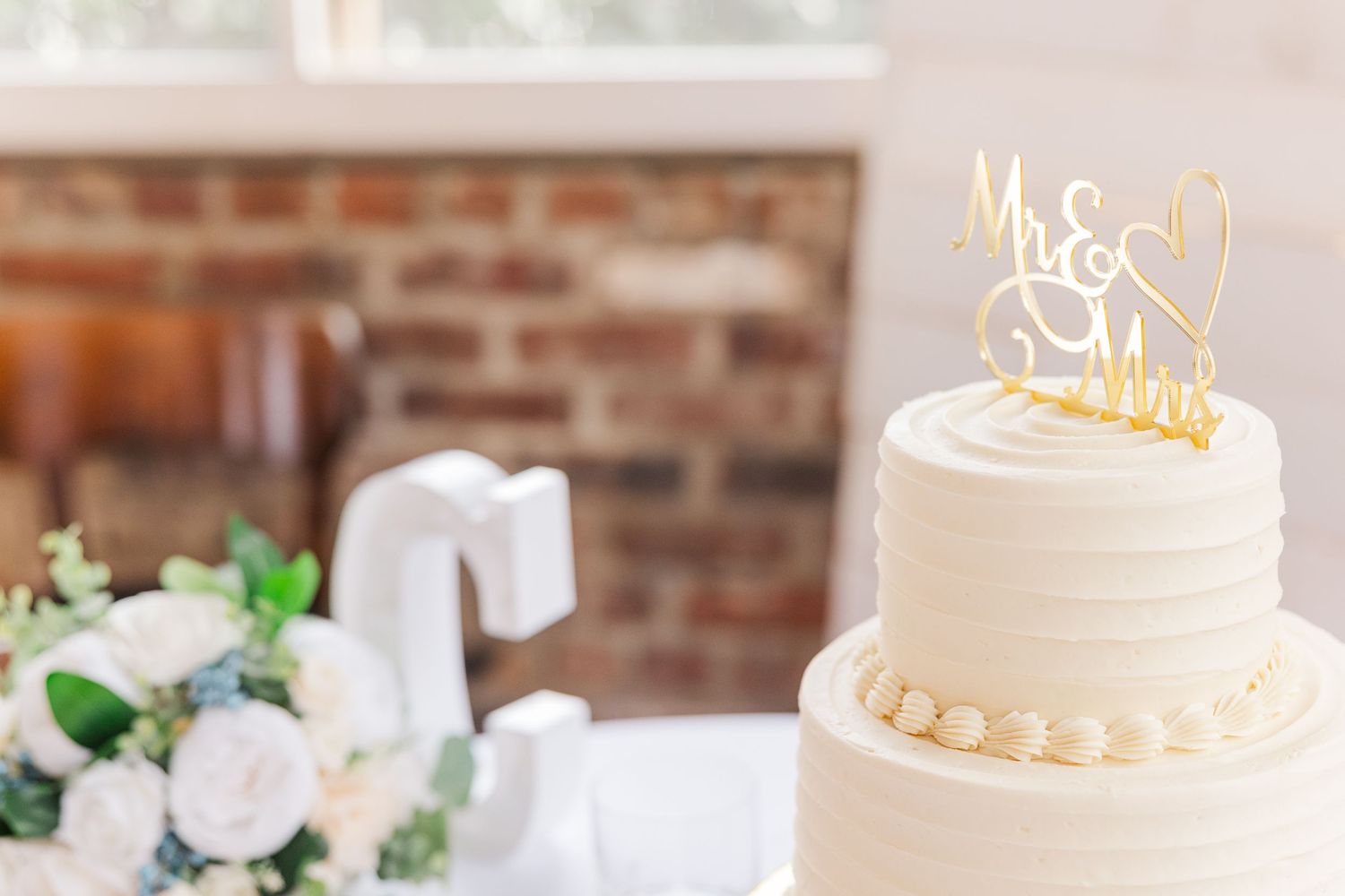 Close-up of white wedding cake with gold metallic topper in rustic venue setting.