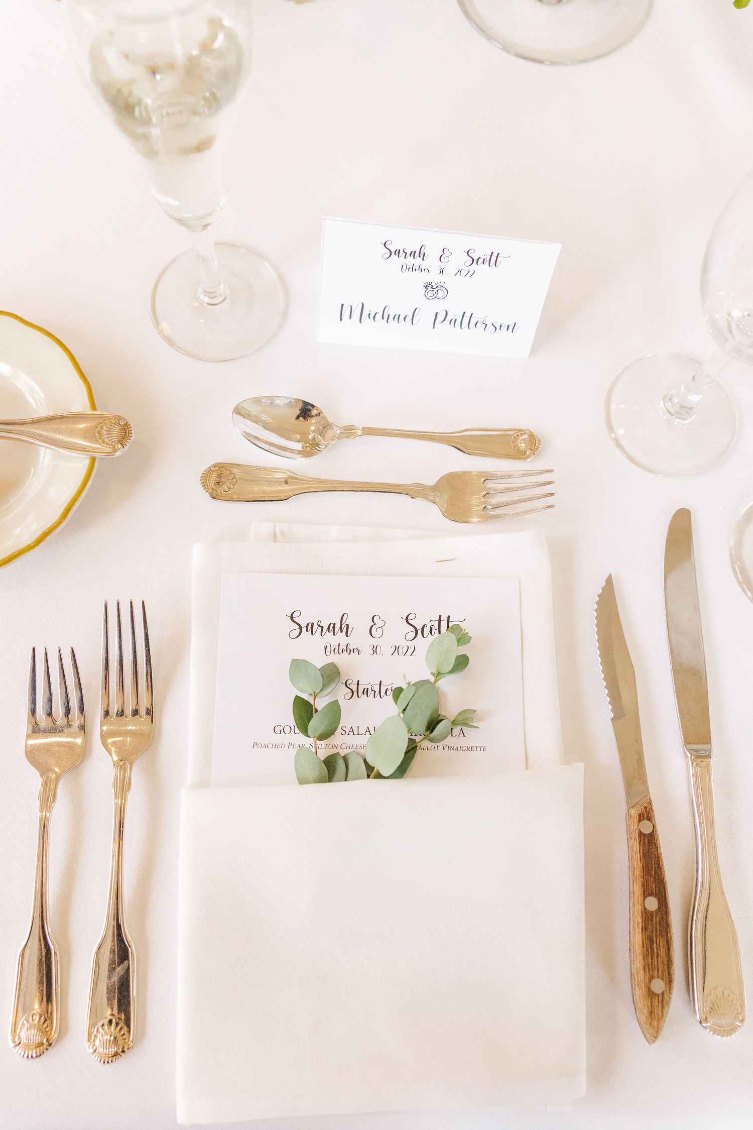 Wedding place setting with gold flatware, name card, and elegant white napkin on white tablecloth.