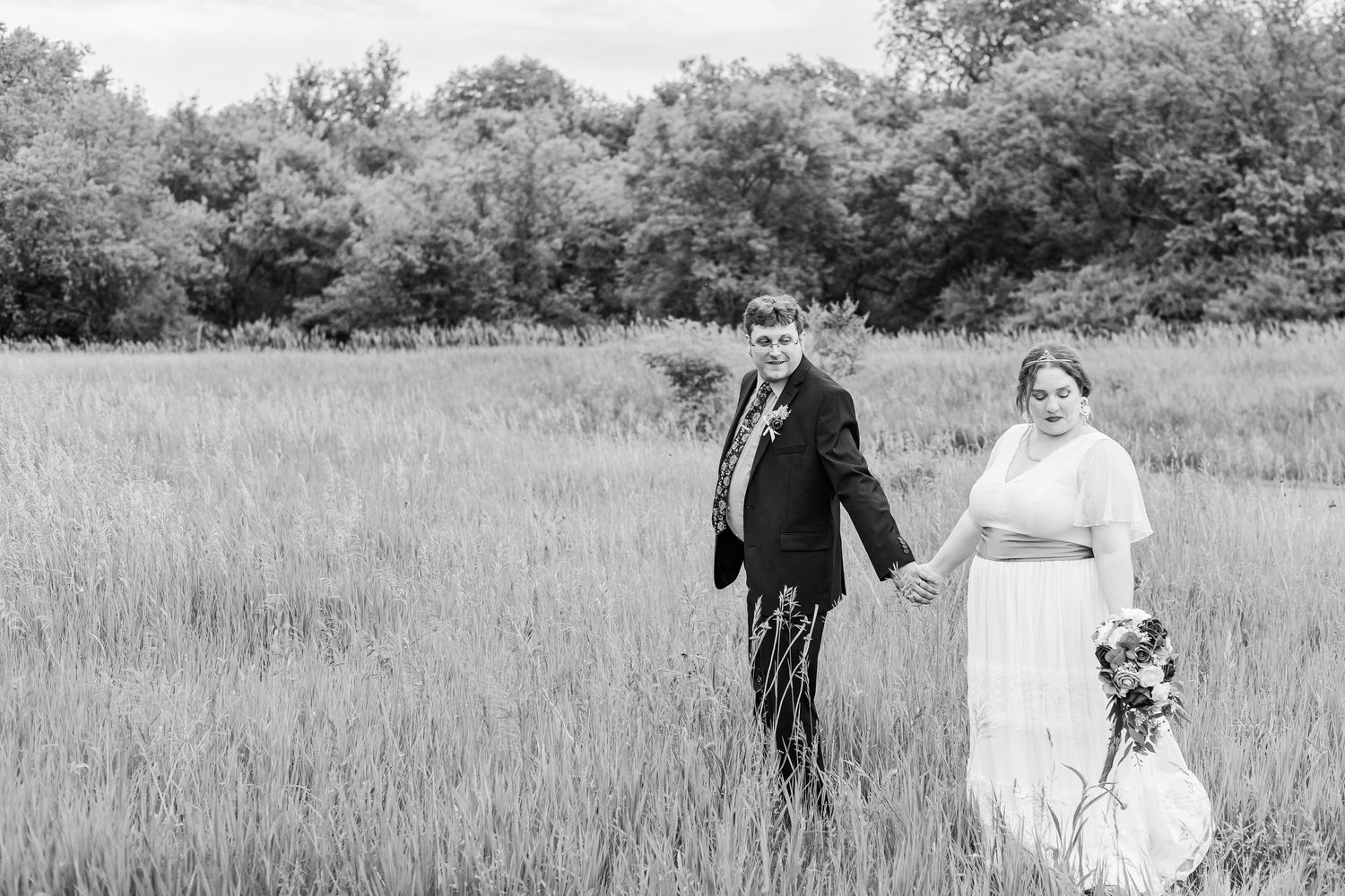 A couple in wedding attire walks through a serene meadow with tall grass and trees in the background.