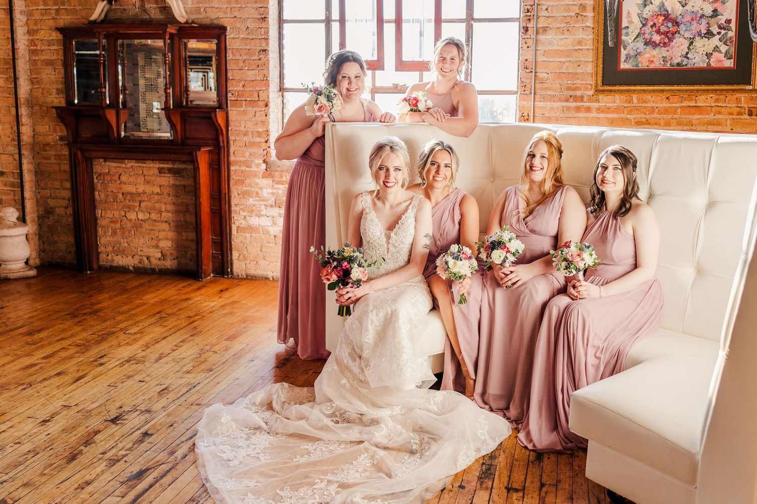 Wedding party seated on white tufted couch wearing dusty rose gowns in rustic venue.