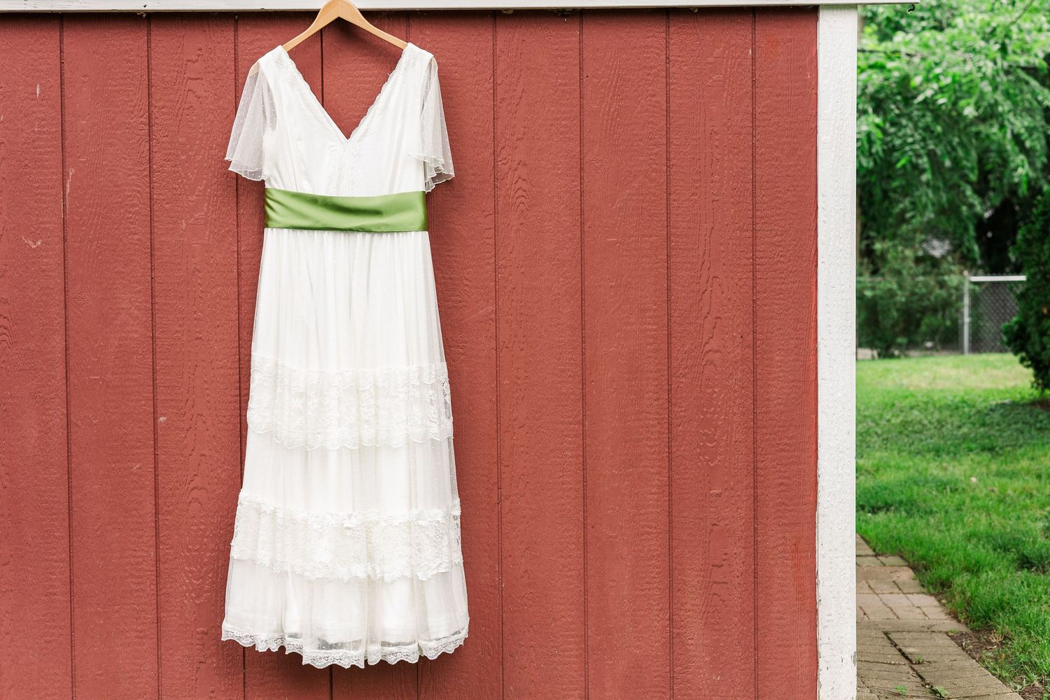 Elegant white wedding gown with green sash displayed on rustic red barn wall.