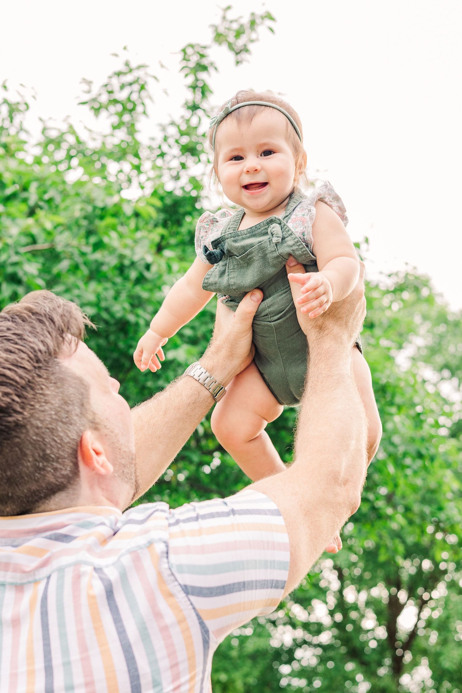 A baby wearing a green romper is lifted high in the air against a backdrop of green leaves.