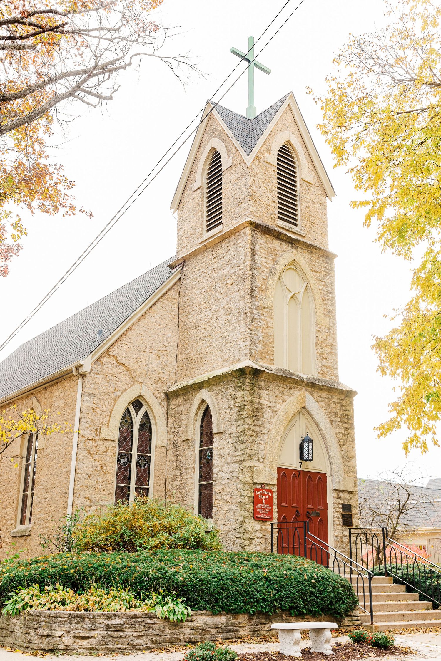 Stone church with Gothic architecture features and red door surrounded by autumn foliage.