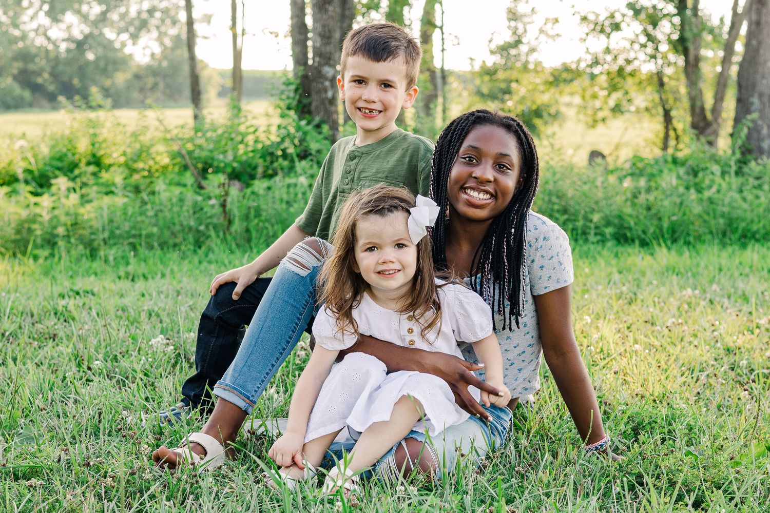 Three people sitting together and smiling on grassy ground in a sunlit outdoor setting.