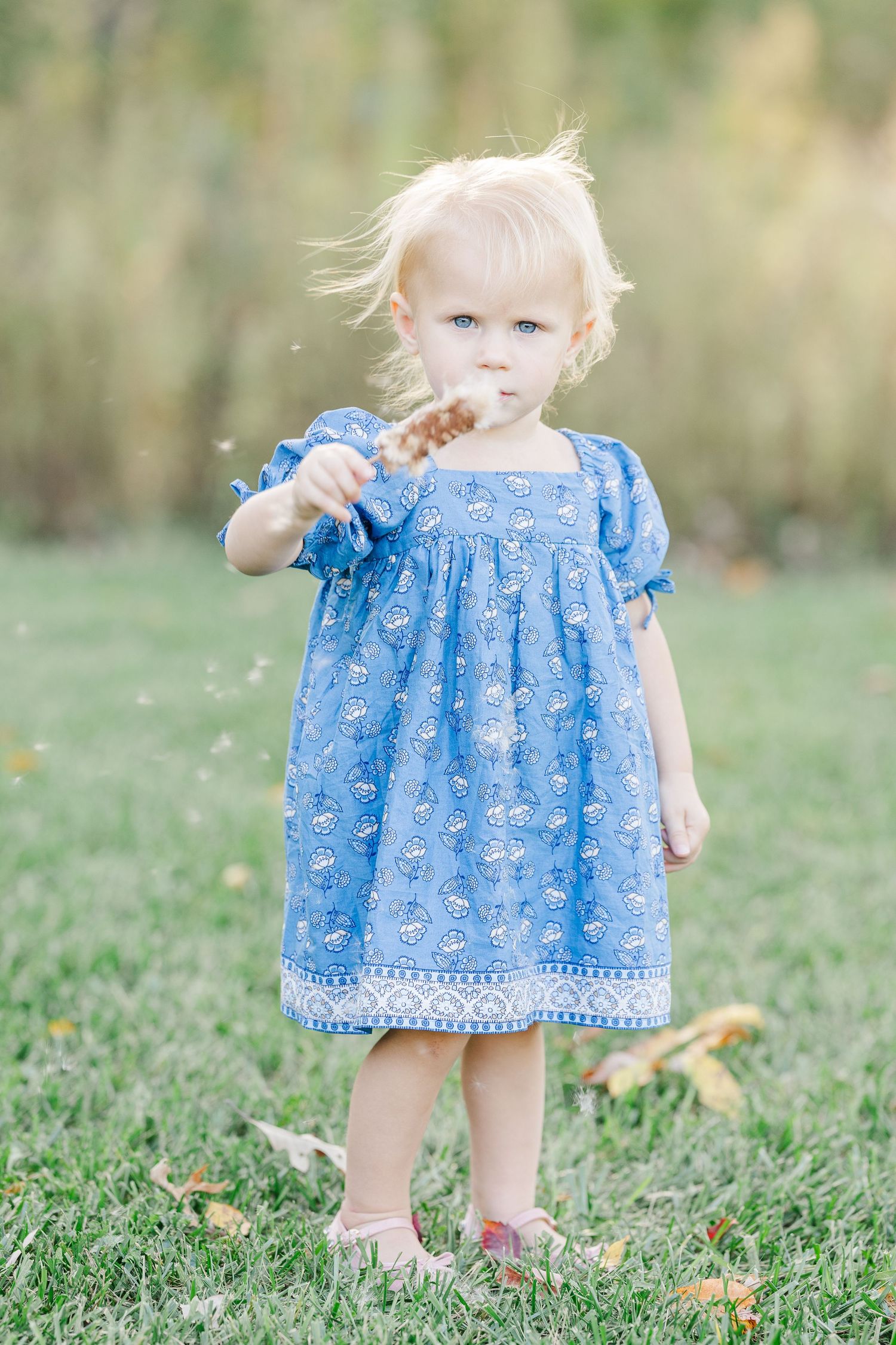 A small child in a blue floral dress twirls and plays in a grassy field on a sunny day.