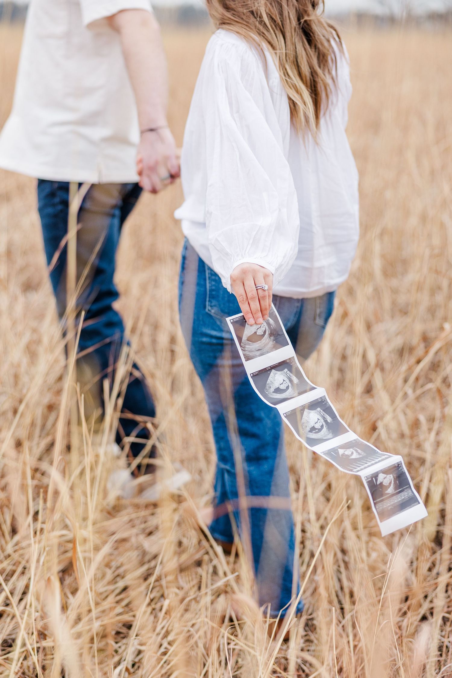 Couple walking through tall grass with ultrasound photos during pregnancy announcement session at Sands Main Street Prairie Cary IL