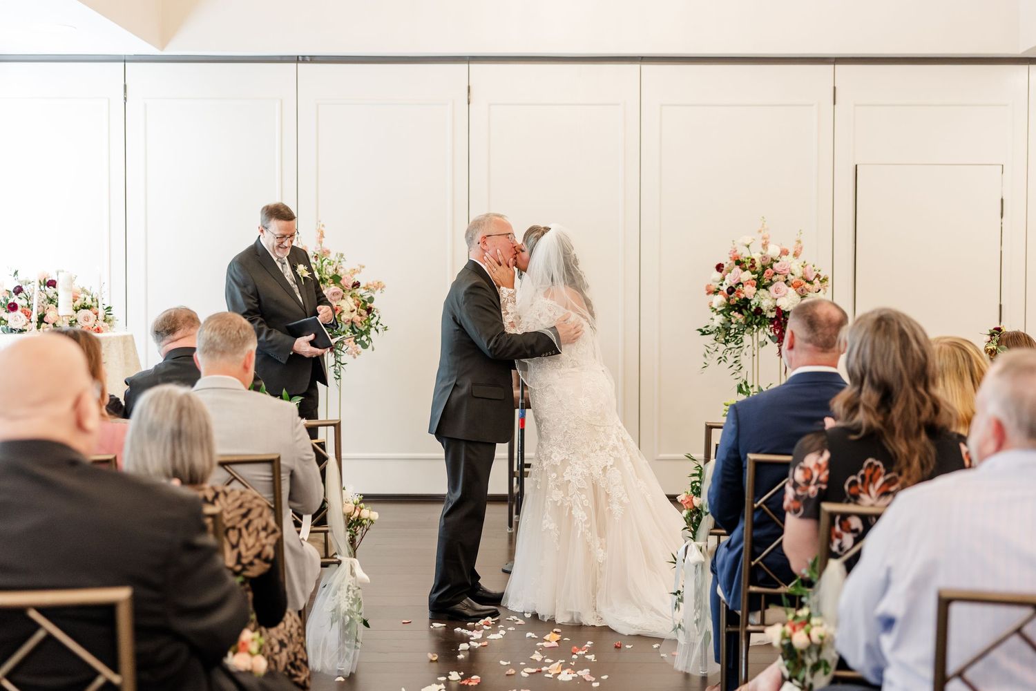 Bride in a lace wedding gown and groom share a kiss at the aisle as guests watch during a elegant indoor ceremony with floral arrangements.
