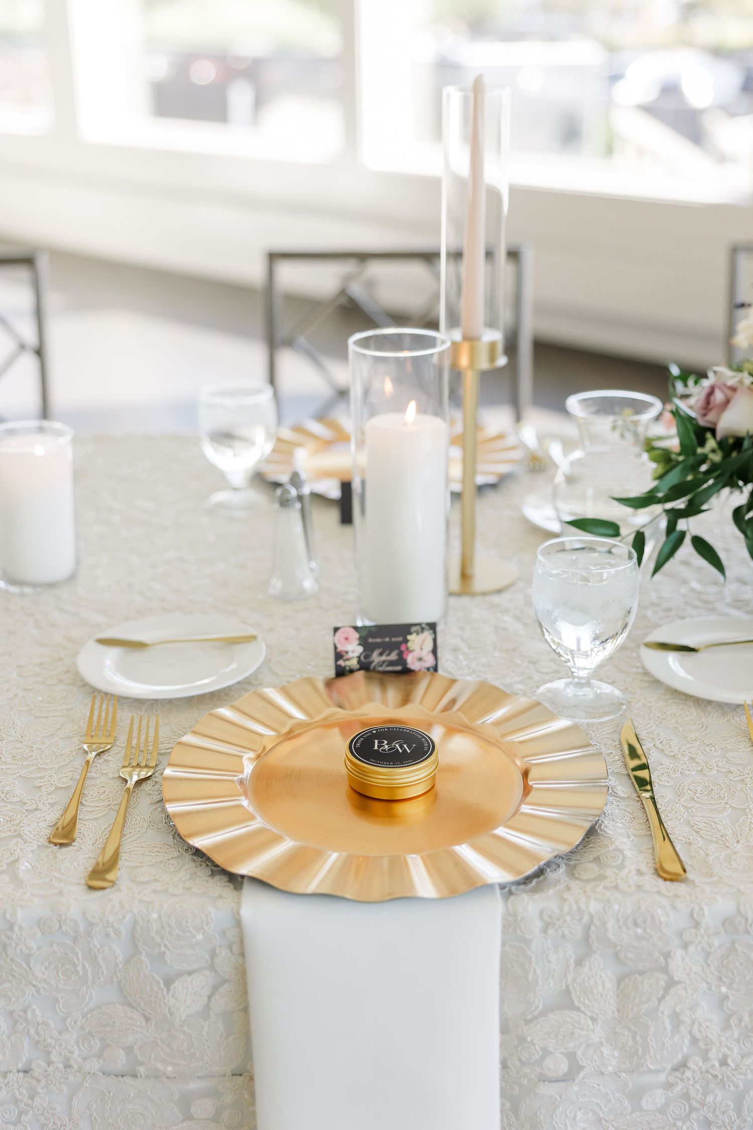 Elegant wedding table setting with a gold charger plate, white candles, gold flatware and floral accents on lace linen.