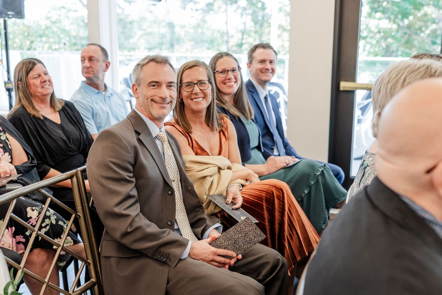 Smiling man in a brown suit sits among attendees during an indoor ceremony, with guests seated behind him near large windows.