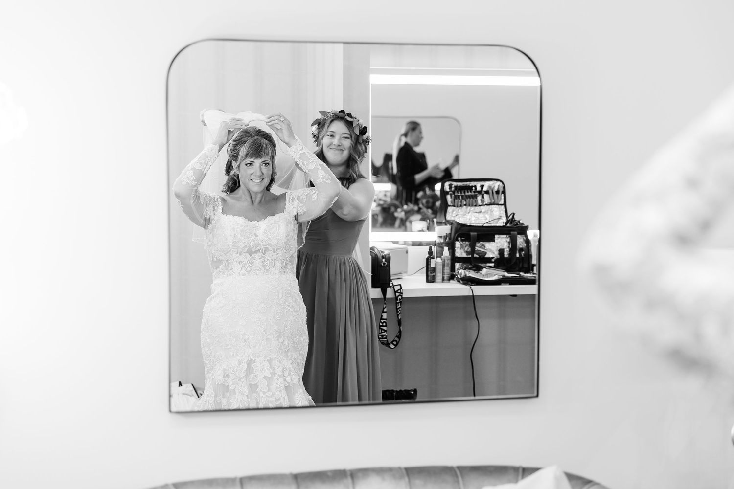 Black and white photo of a bride in a lace gown adjusting her veil in a mirror as a bridesmaid stands behind her getting ready.