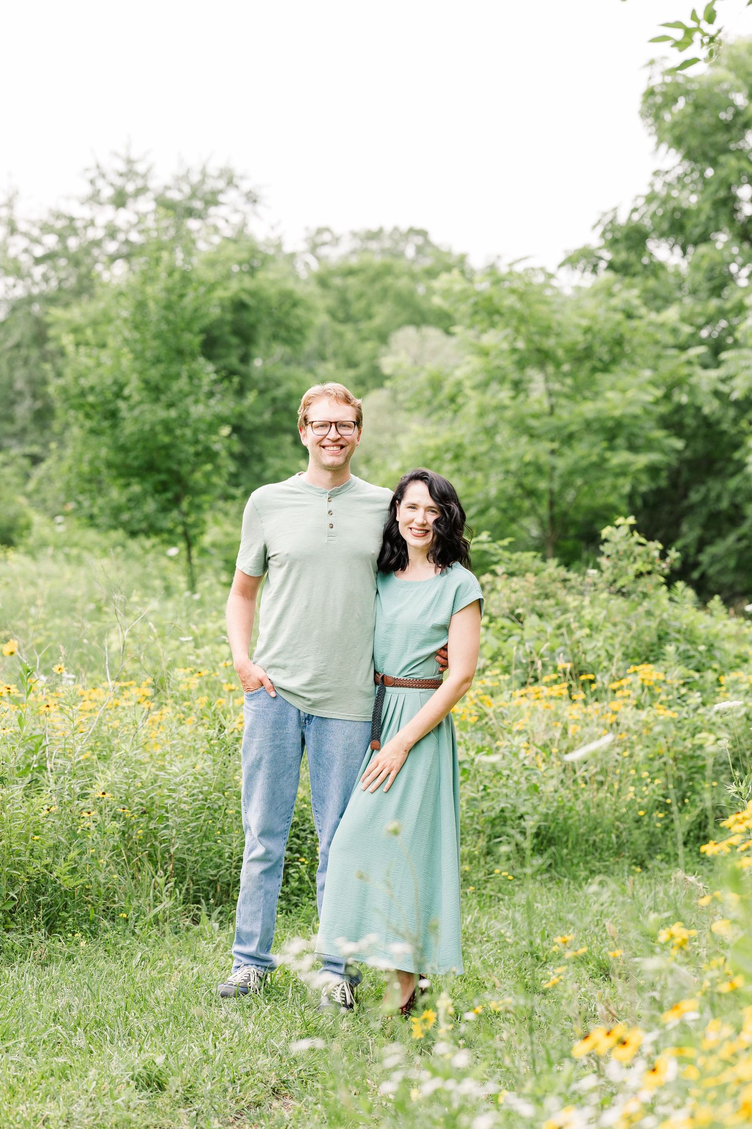 A smiling couple stands together in a lush green meadow with yellow wildflowers, woman in teal dress, man in green shirt.