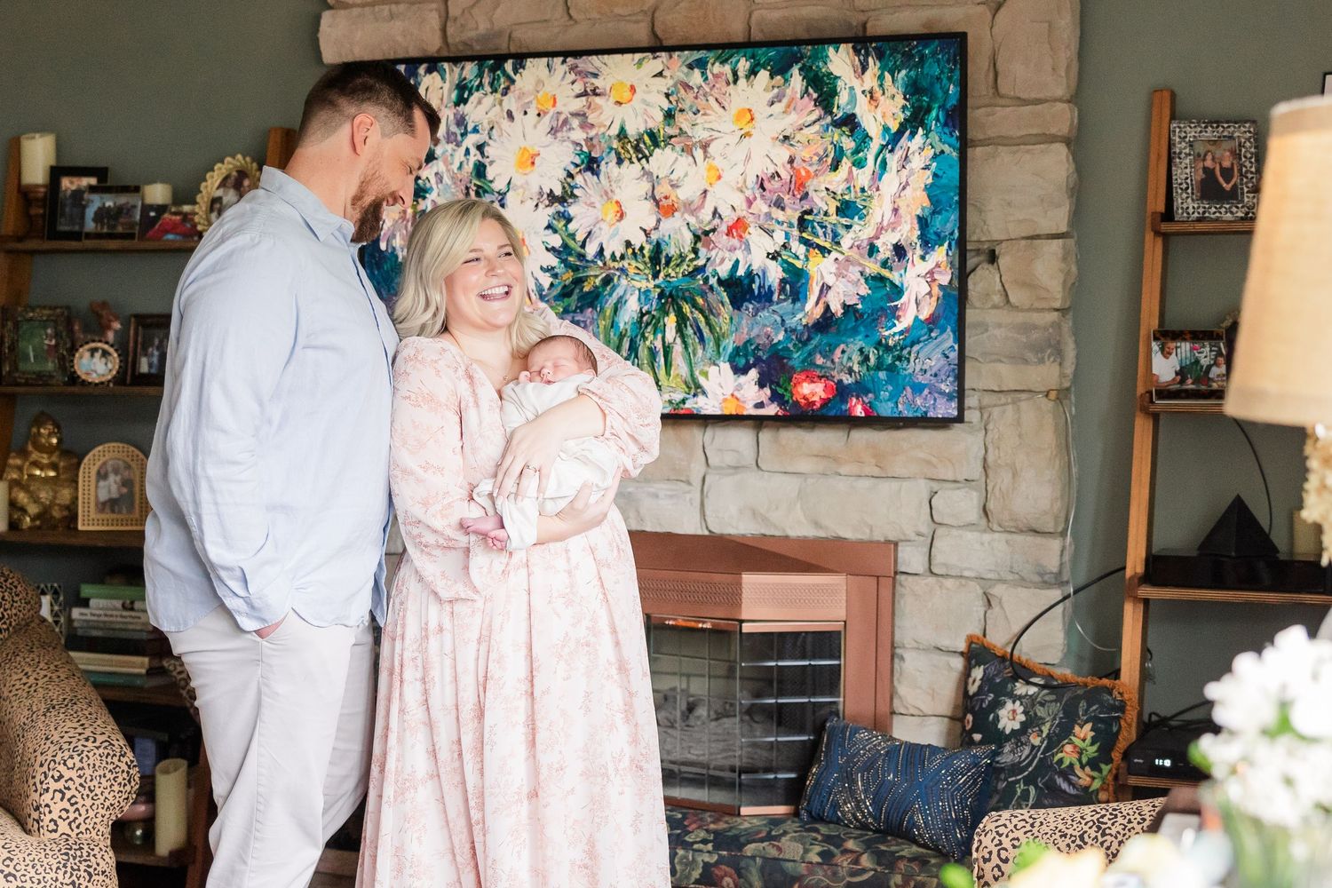 New parents stand together holding their newborn baby in a cozy living room with a floral painting above the fireplace.
