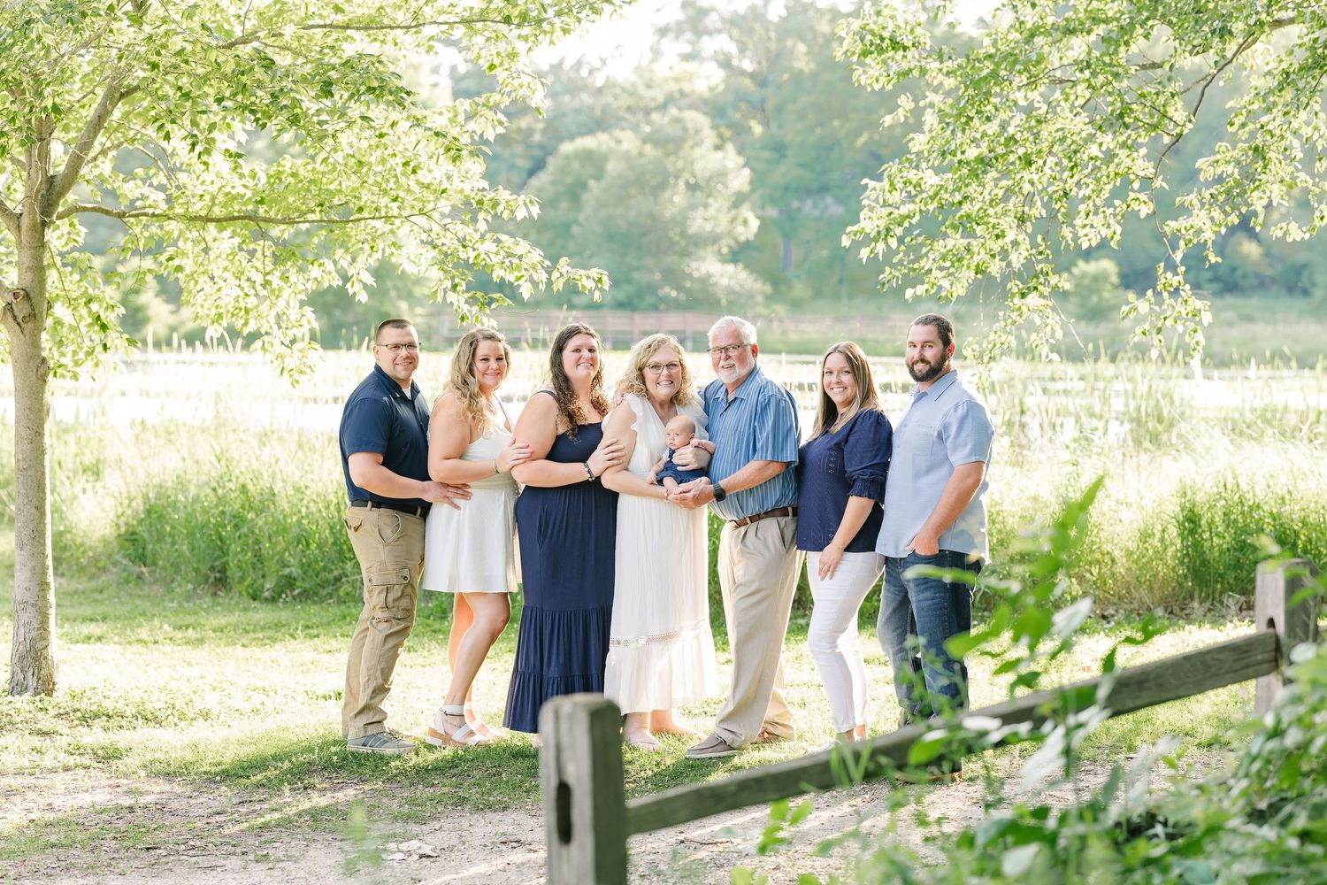 Summer extended family portrait at Veterans Happy family of seven smiling together outdoors near a wooden fence with green trees and fields.