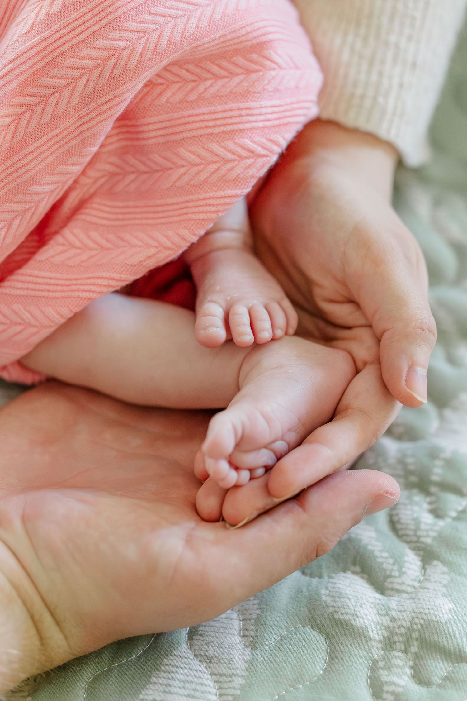 Two pairs of adult hands gently cradling tiny newborn baby feet wrapped in a soft pink blanket.