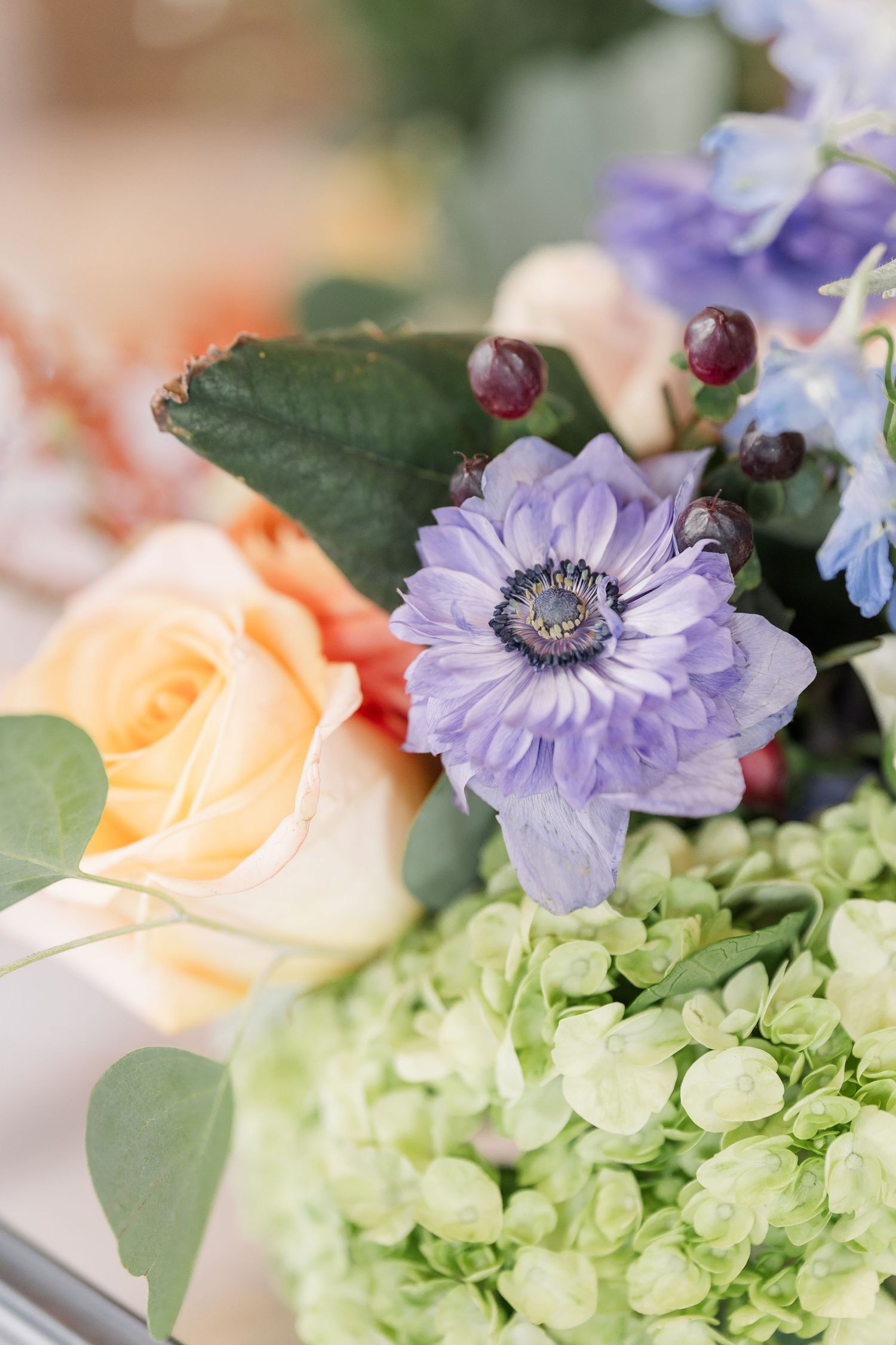 Close-up of a colorful floral arrangement featuring a purple anemone, peach rose, and green hydrangea.