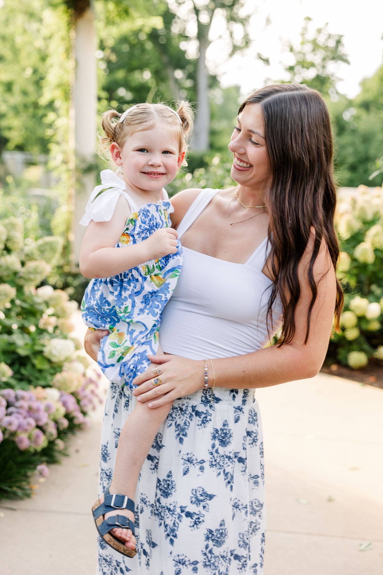 A smiling brunette woman in white top and floral skirt holds a happy toddler girl in a blue floral dress outdoors.