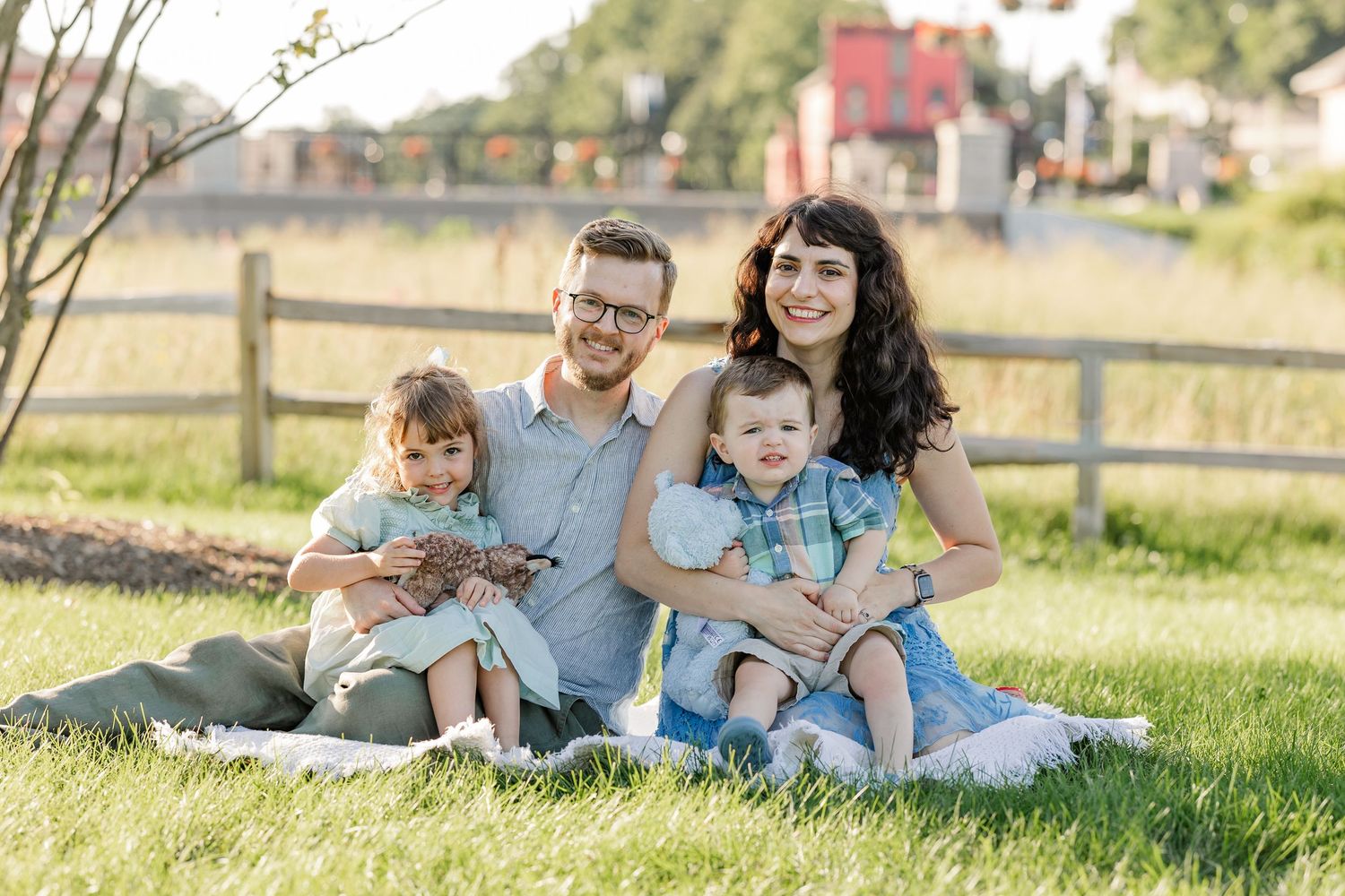 Happy family of four sitting on grass, parents with two young children smiling outdoors near a wooden fence.