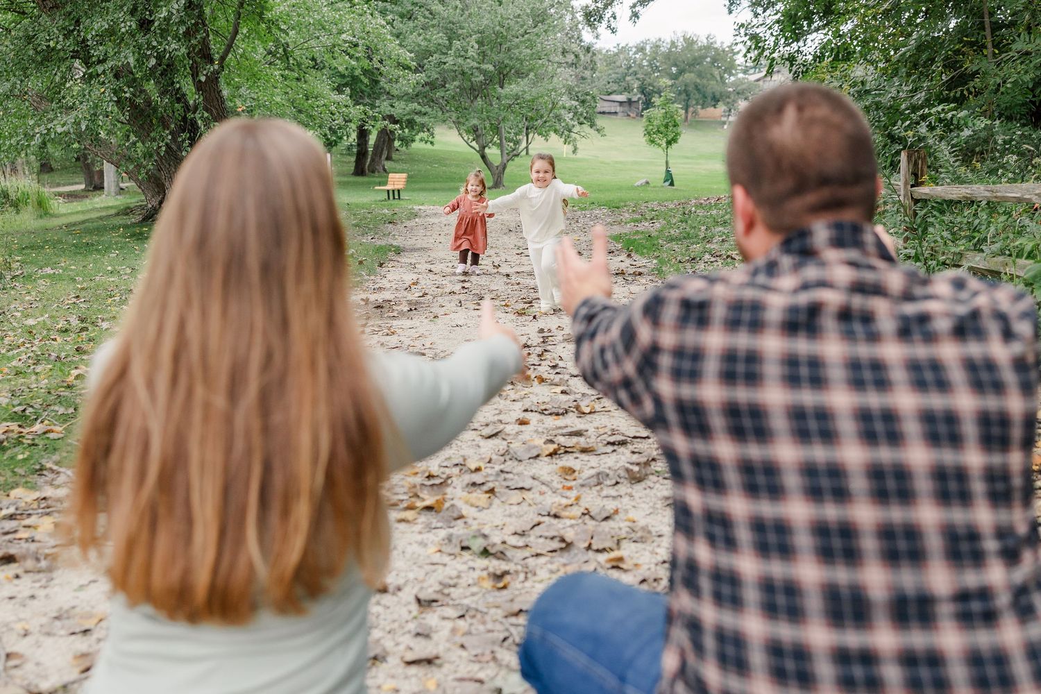 Parents waving to their two young children running toward them on a park path surrounded by green trees.