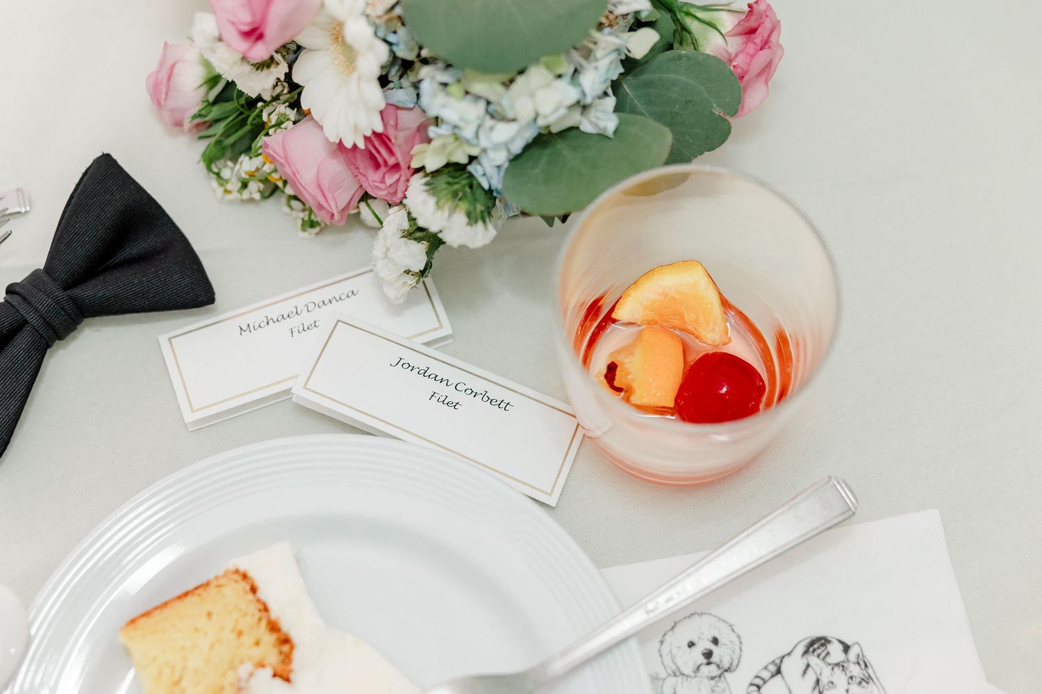 Wedding place setting with cocktail, cake slice, bow tie, flowers, and name cards on table.