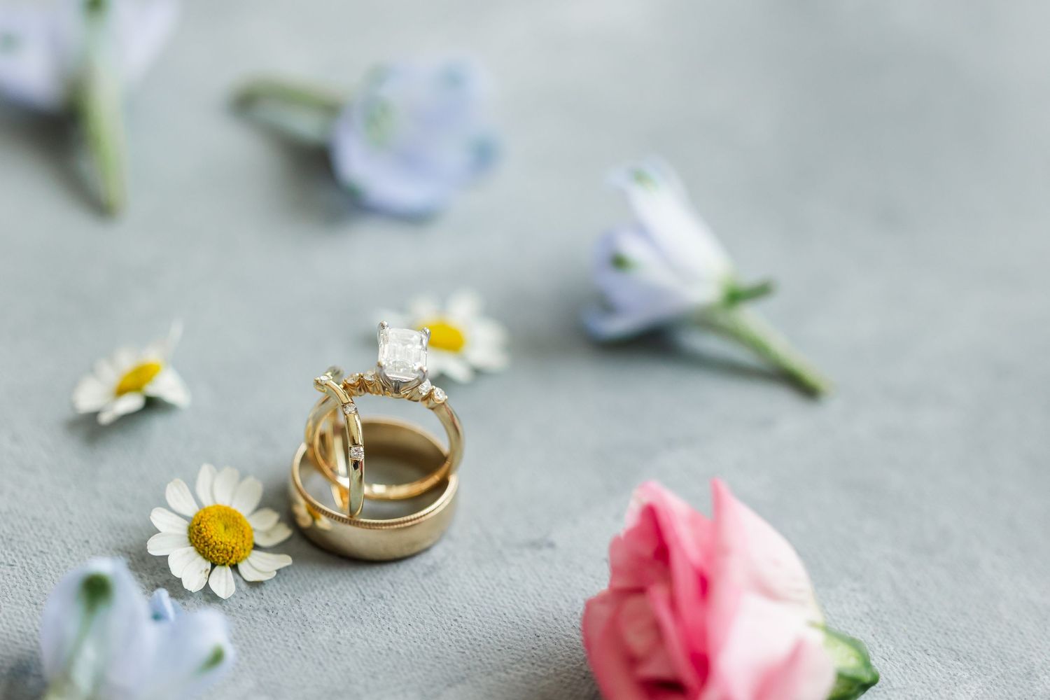 Gold wedding rings with diamond surrounded by delicate daisies, pink roses, and blue flowers on gray background.