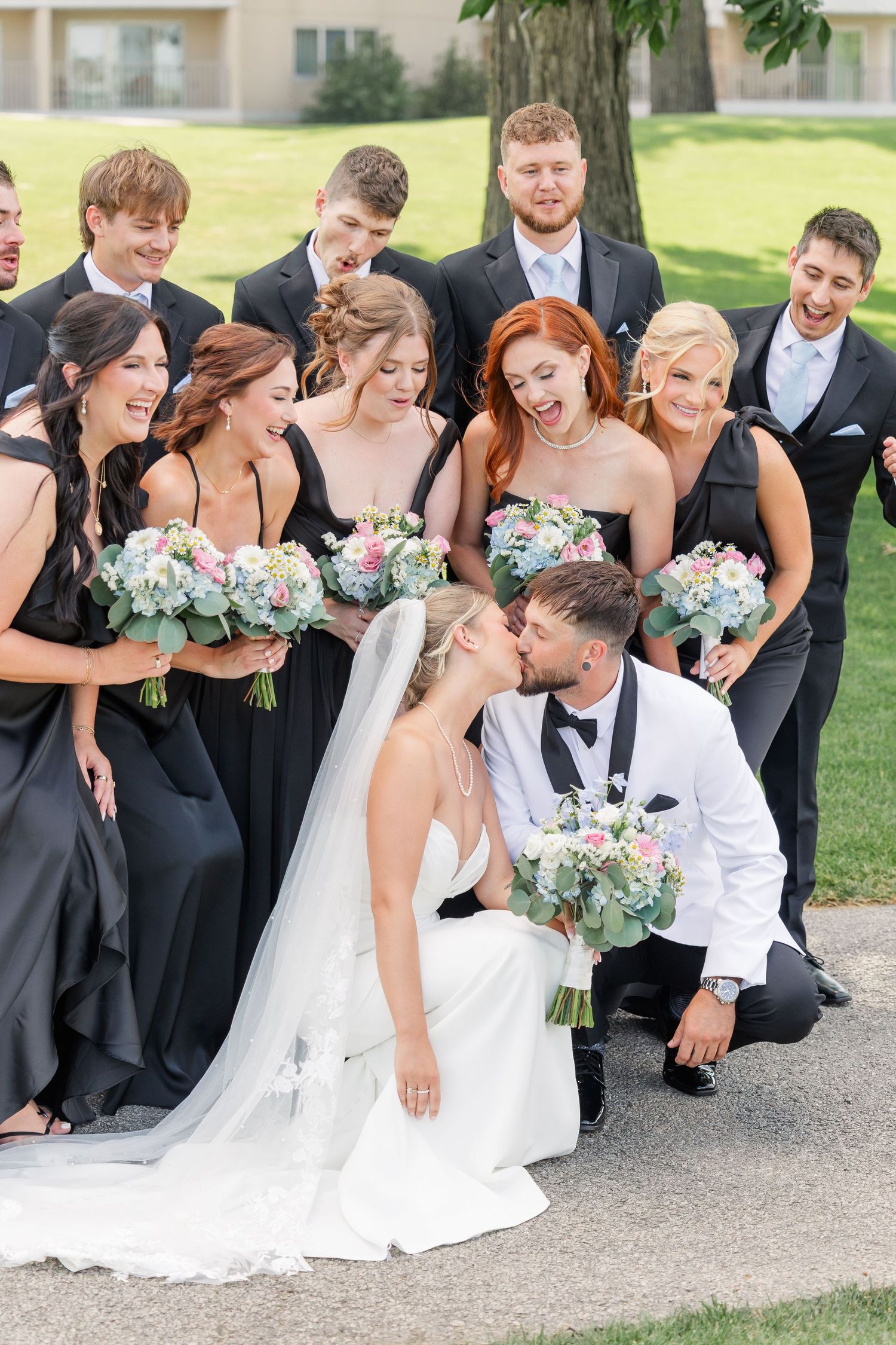 Wedding party group photo with bride and groom kissing, bridesmaids in black dresses holding floral bouquets.