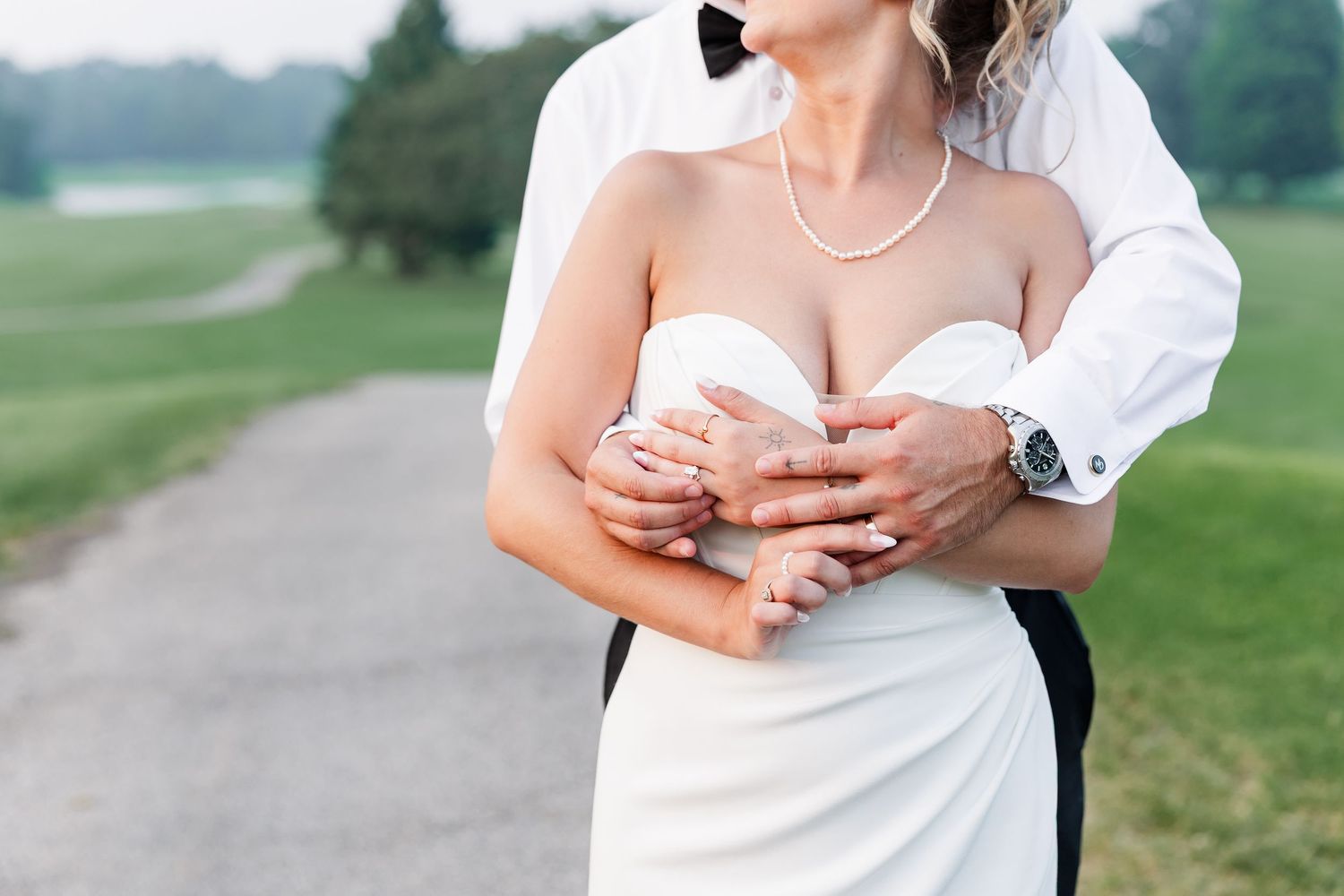 Bride in strapless white gown wearing pearl necklace embraced by groom in black suit on golf course path.