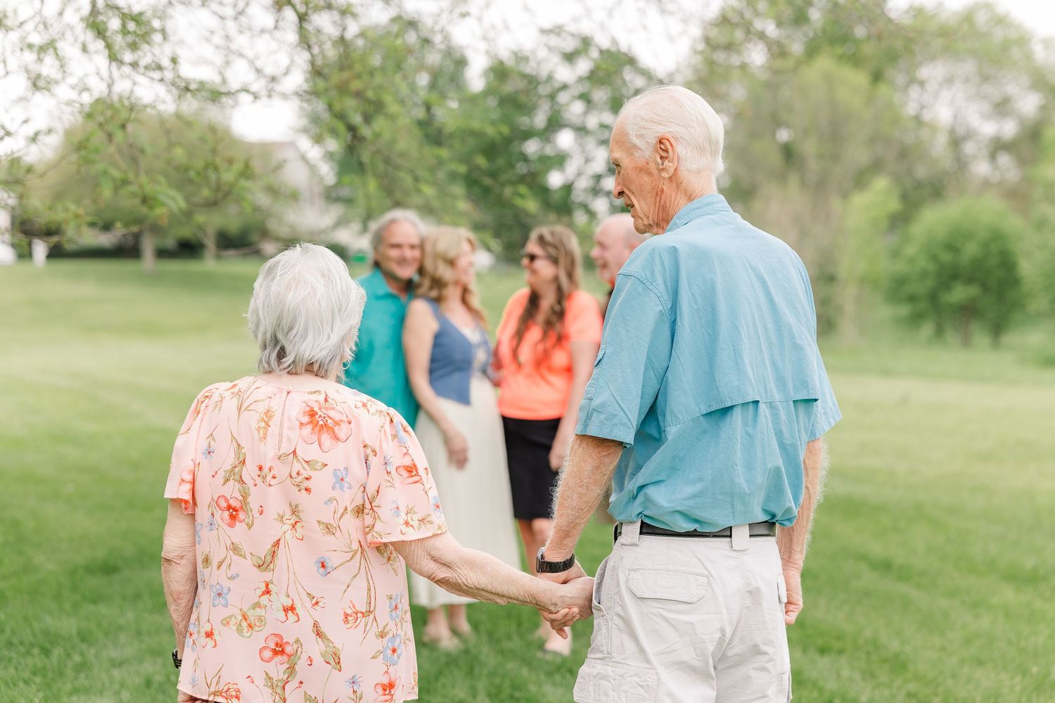 Elderly couple holds hands walking toward family group in lush green park during outdoor gathering.
