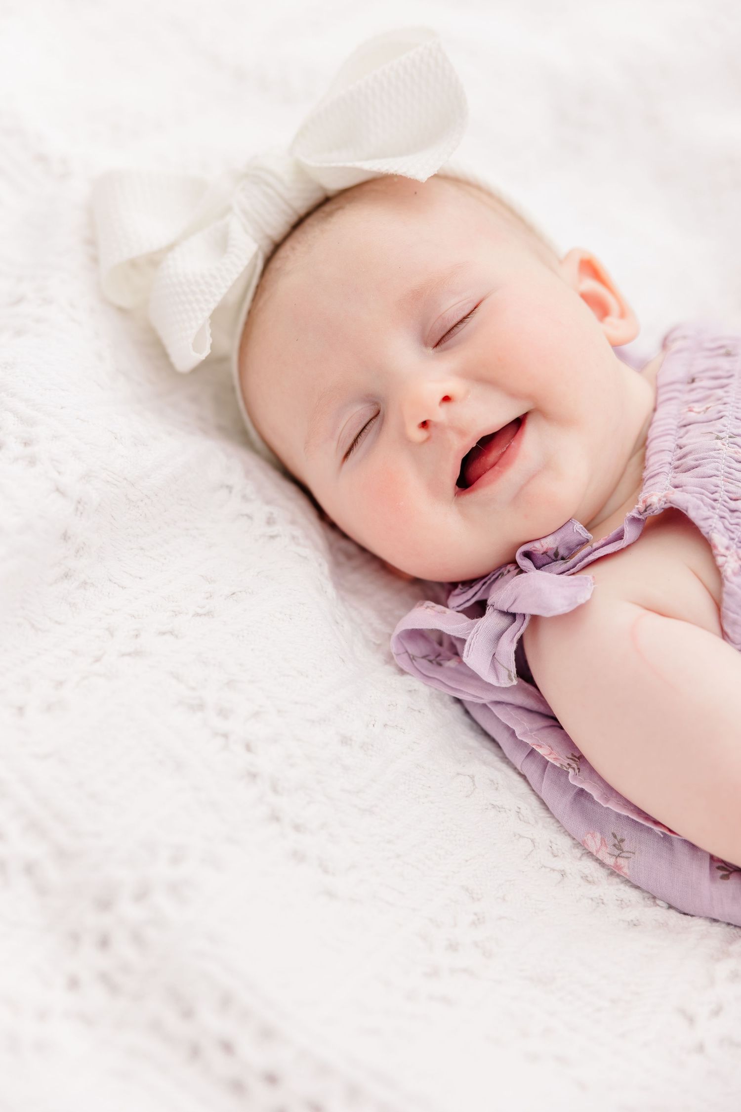 Adorable smiling baby in lavender outfit and white bow headband lying on a white knit blanket.